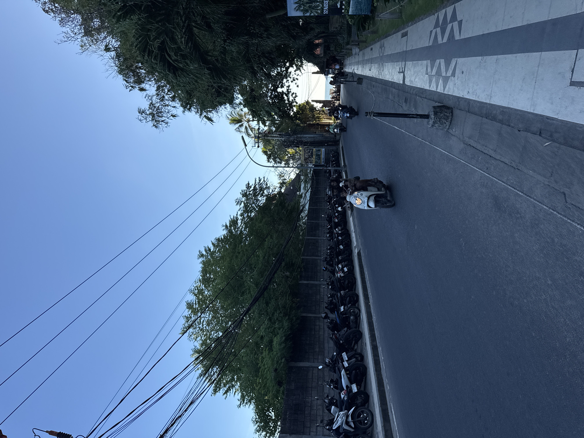 The image depicts a street scene in Kuta, Indonesia. The view is oriented lengthwise along the street, which is lined with parked motorbikes on one side and a pedestrian walkway on the other. Trees and utility lines are visible along the motorbike side of the street. Two motorbikes with riders are visible moving along the road, moving towards the ocean in the distance. The pedestrian walkway is patterned with decorative markings. The sky is clear and blue.