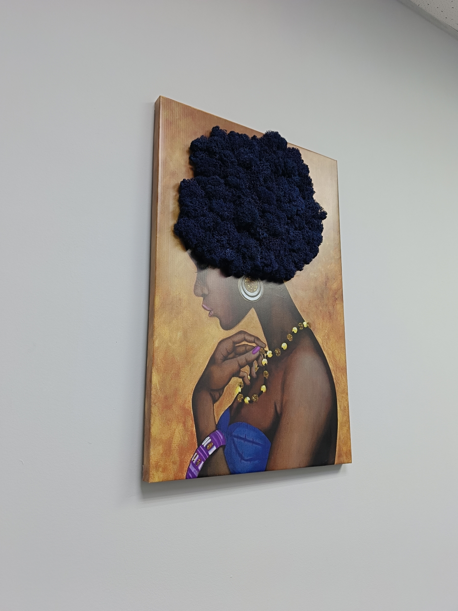 An eye-level, slightly angled photograph captures a distinctive mixed-media artwork hanging on a plain, light-colored wall. The artwork depicts the profile of a woman, facing towards the left side of the frame. Her skin is rendered in warm brown tones, and she wears a vivid blue garment, possibly a strapless top or dress.
A prominent feature of the artwork is the woman's hair, represented by a voluminous, dark blue, textured, three-dimensional material that protrudes from the canvas, giving it an afro-like appearance. She is adorned with a large, intricate circular earring and a necklace composed of yellow and gold beads. Her right hand, with purple-painted fingernails, is visible near her neck, appearing to gently touch or adjust the necklace. A purple and white striped bracelet or arm cuff is visible on her left arm.
The painted background behind the woman is a warm, textured golden-brown, creating a rich contrast with her features and accessories. The artwork is framed with a simple, light brown wooden edge. The lighting appears to be ambient indoor light, and no specific time of day can be determined. The wall itself is a uniform light grey or off-white. There is no visible text in the image. This media file originates from Chaikovsky, Russia.
