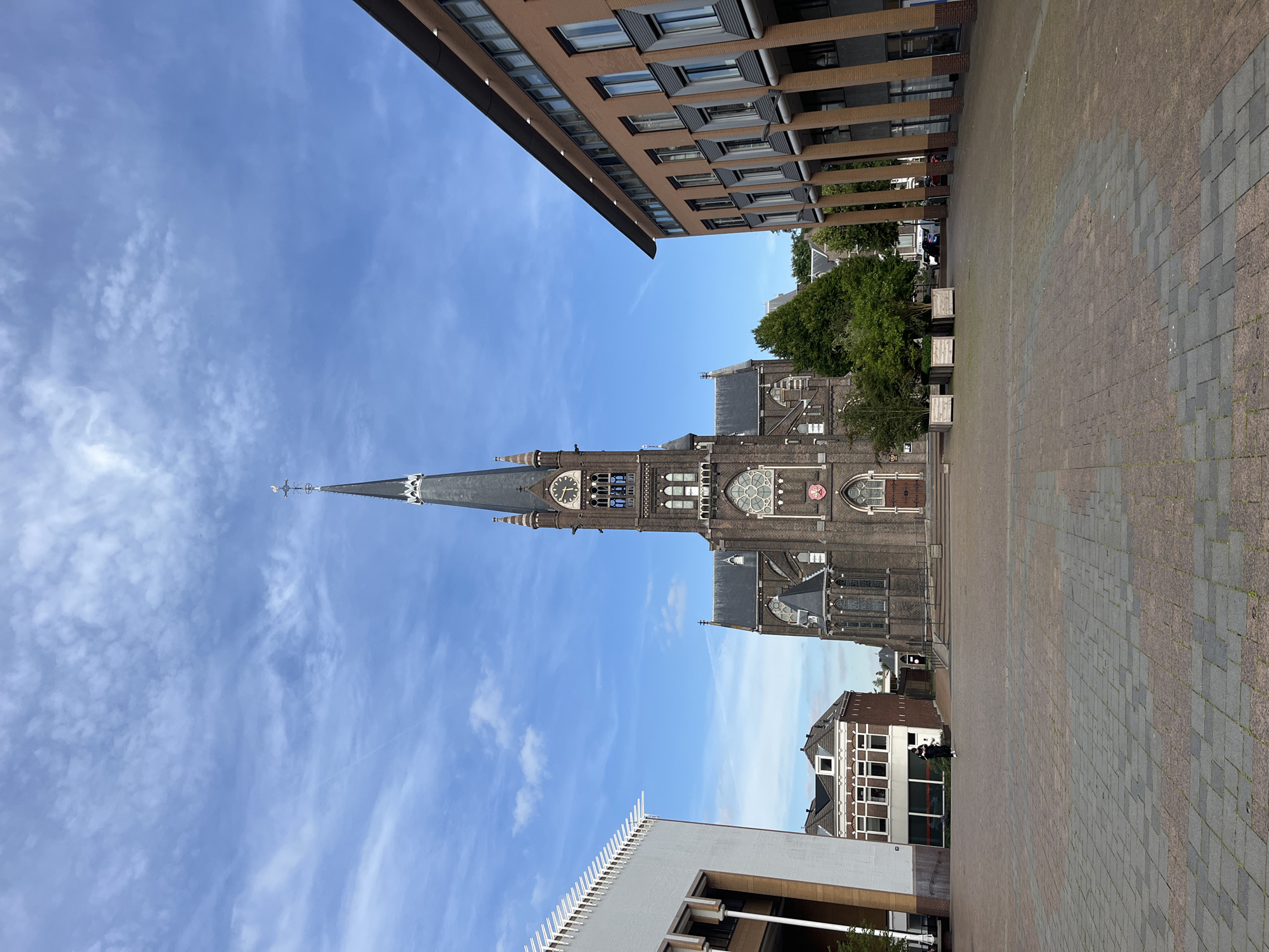 An eye-level, medium-wide shot captures a large, dark brick church with a tall steeple dominating the center of the frame. The church features intricate architectural details, including arched windows, a rose window, and a clock face on the steeple. To the left of the church, a modern white building with vertical lines stands prominently. To the right, a contemporary brick building with columns lines the edge of the square. The ground in the foreground is paved with interlocking stones in a patterned arrangement of reddish-brown and gray. A lone figure, dressed in dark clothing, walks across the square towards the left side of the frame, with their back to the camera. The sky above is a bright blue, adorned with scattered white clouds. The environment appears to be an urban square in the city of Unknown, Unknown.