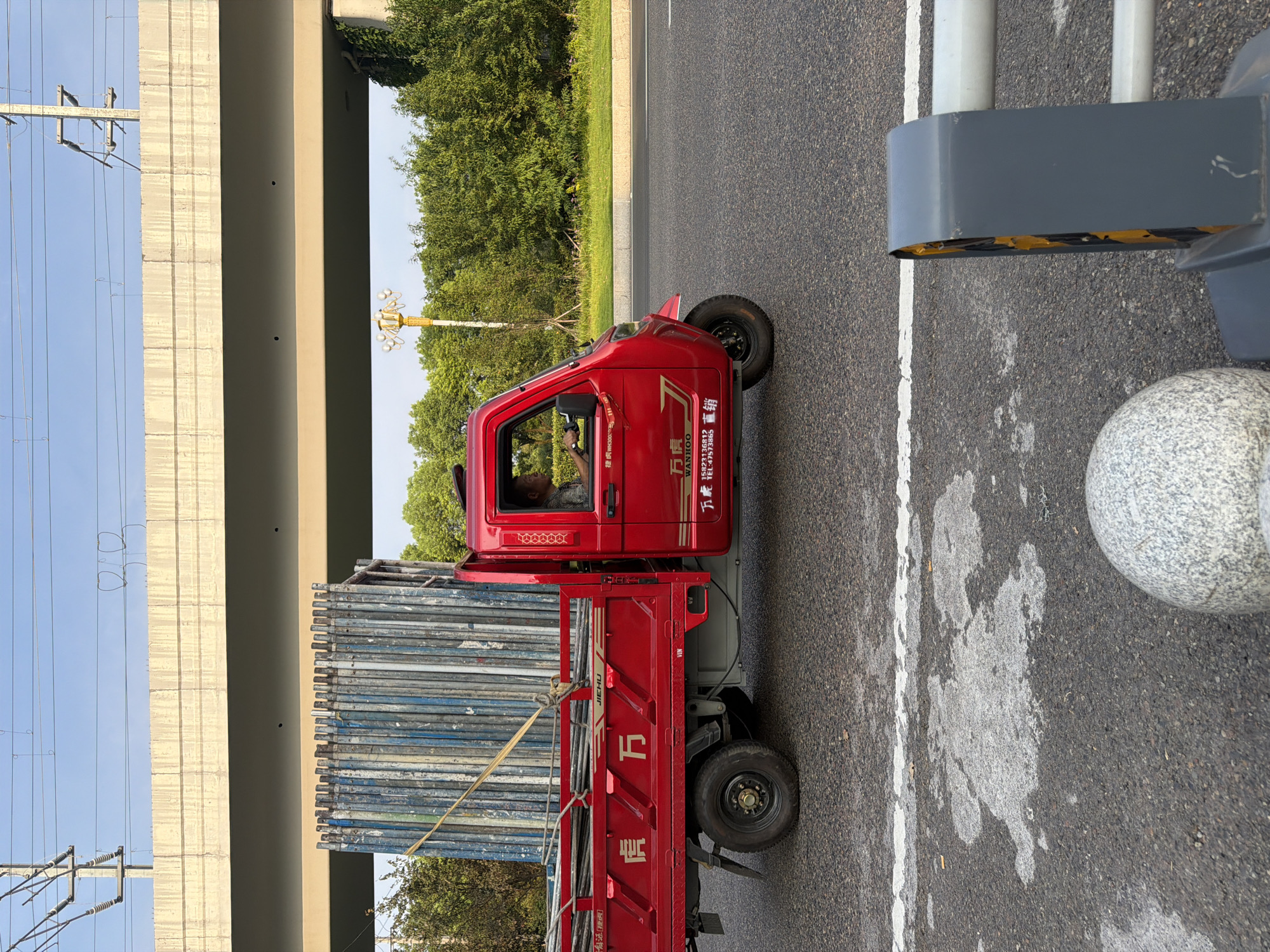 A side profile of a bright red, three-wheeled truck is captured on a paved road, likely in Jiangjin, China, as indicated by the presence of Chinese characters on the truck. The truck is transporting a large load of metallic scaffolding poles, which are bound by a yellow strap. A male driver, wearing a patterned short-sleeved shirt, is visible inside the truck's cabin, his right hand on the steering wheel. The truck has Chinese characters on its side, including "万虎" and "JIEHU." The road appears to be an elevated highway or a road passing under an overpass, evidenced by the concrete structure overhead. The environment is characterized by lush green trees lining the road and a clear sky with overhead power lines. The foreground of the image shows a portion of the road with white markings, and a gray barrier with yellow and black elements. A spherical, textured object, possibly a bollard or a decorative element, is partially visible in the bottom left corner.