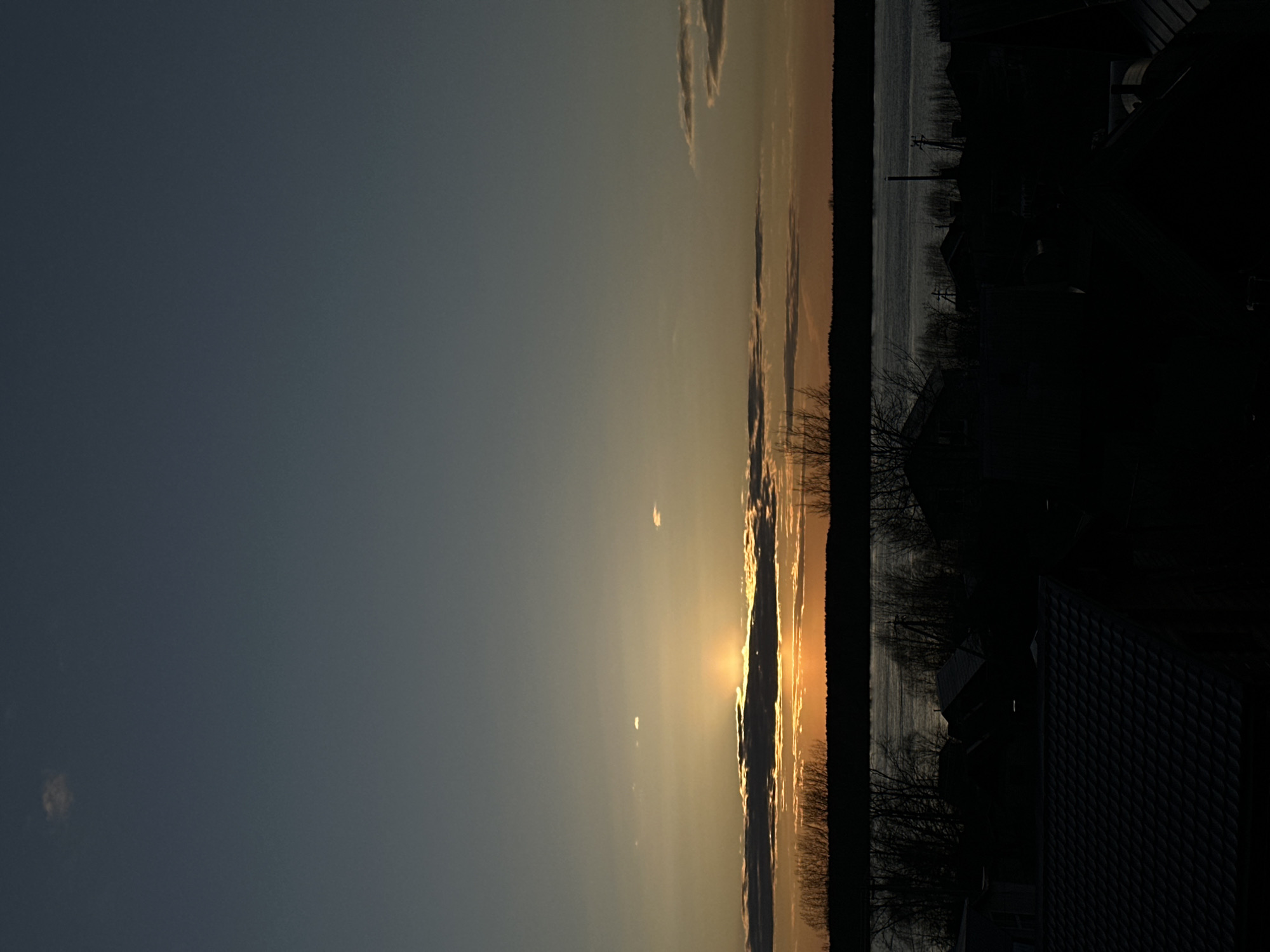 The image depicts a sunset over a body of water, possibly the Kama River, from Chaikovsky, Russia. The sun is partially obscured by clouds, casting a golden light over the horizon and the water. The foreground consists of silhouetted trees and rooftops of houses, suggesting an urban or suburban location. The sky transitions from a dark blue to a lighter orange and yellow near the horizon. A narrow strip of dark trees forms a distinct line between the sky and the water. The overall scene evokes a sense of tranquility and evening atmosphere.