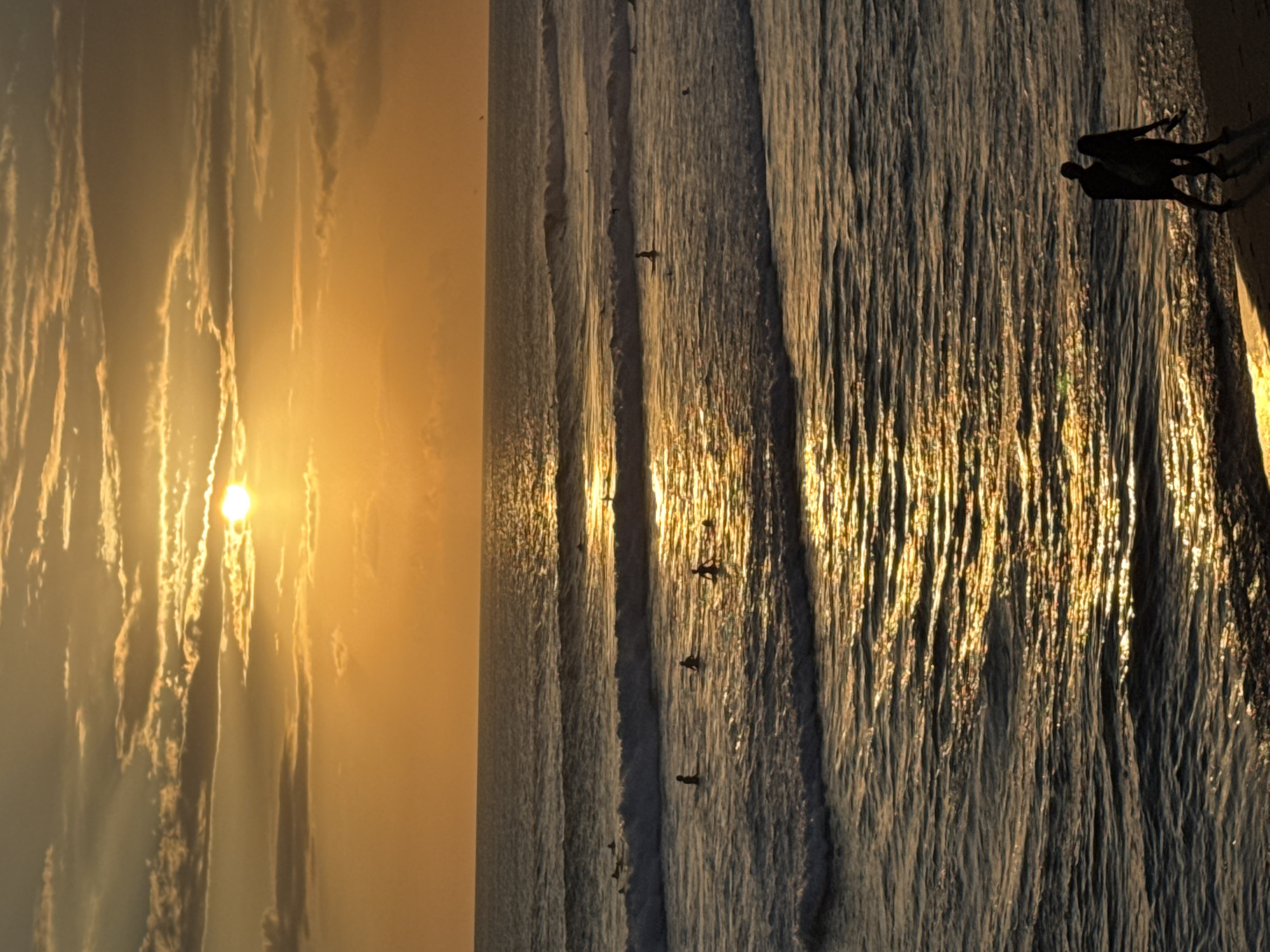 The image depicts a sunset scene at a beach in North Kuta, Indonesia. The sun is low in the sky, casting a golden glow over the clouds and water. There are people, presumably surfers, visible in the water. Two people are silhouetted on the sand, apparently interacting. The sea has a textured appearance, reflecting the sunlight.
