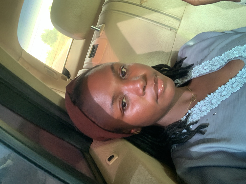 A young Black female is seated in the rear passenger compartment of a vehicle in Gadam, Nigeria. She occupies the central foreground, looking directly at the camera with a neutral expression. She wears a dark reddish-brown headwrap covering the crown of her head, with dark dreadlocks or braids visible underneath. Her attire consists of a light blue or grey garment with intricate light blue lace trim along the neckline. A gold-colored necklace with a small circular pendant adorns her neck, and a metallic stud is present in her left nostril. The vehicle's interior features tan or beige upholstered seats and headrests. Through the rear window, a sunlit outdoor environment is visible, characterized by a light blue sky, sparse green foliage or trees, and a dirt road or barren ground in the distance, suggesting a rural or semi-rural setting under clear daytime conditions. The subject is static, posing for a photograph.