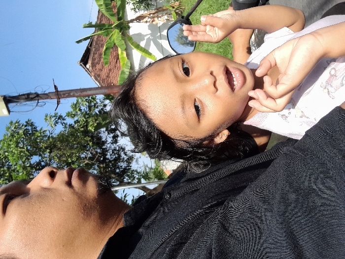 A man in a black shirt holds a child in his arms while on a motorcycle. The child faces the camera, smiling with an open mouth. A motorcycle mirror is visible behind the child. In the background, there's a white building with a brown roof, banana trees, a power pole, and other greenery against a blue sky. The photo is taken from a low angle, slightly rotated.