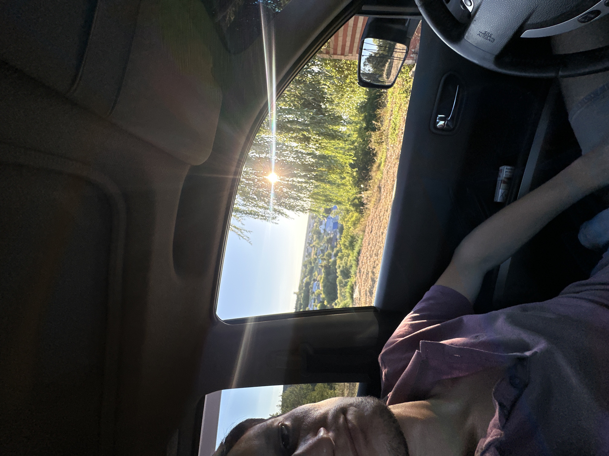 A man is sitting in the passenger seat of a car, looking towards the camera. The man has short, dark hair and is wearing a purple polo shirt. The car's interior is dark, with the exception of the sunlight streaming in through the windows. Outside the car, a rural landscape is visible, with rolling hills, trees, and a village with blue-roofed houses. The sun is shining brightly, casting a lens flare across the image. The side mirror of the car reflects a brick building and trees. A pack of cigarettes is visible in the door pocket. The scene appears to be in or near Makiv, Ukraine, suggested by the rural setting and architecture.