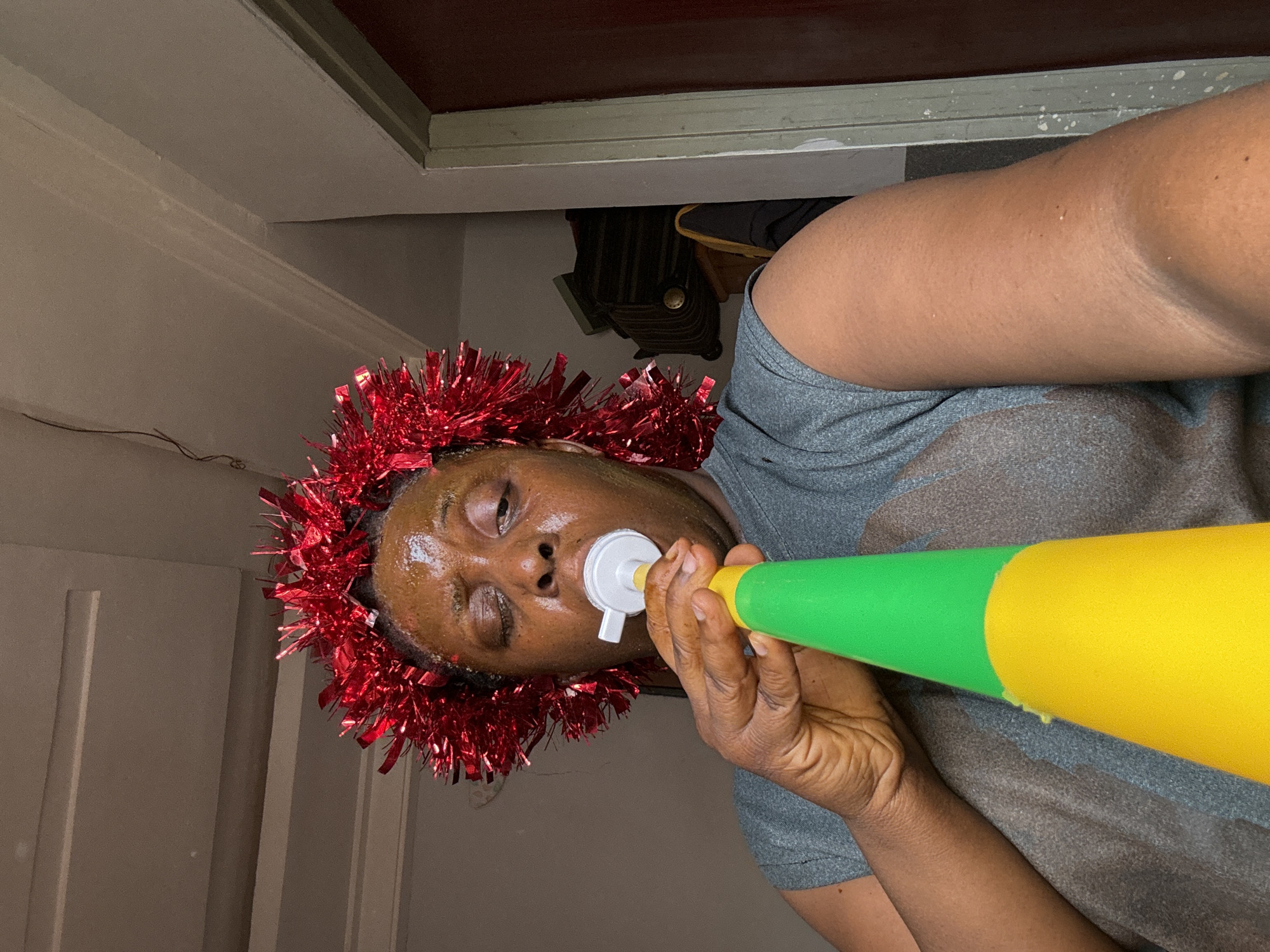 An individual with dark skin wearing a gray shirt and a red tinsel garland is holding a yellow and green party horn in their mouth. The person appears to be indoors, near a door. A backpack is visible behind the individual and door. The photograph appears to be taken at an angle. The scene does not offer enough information to indicate that this image was taken in Sabon Tasha, Nigeria.