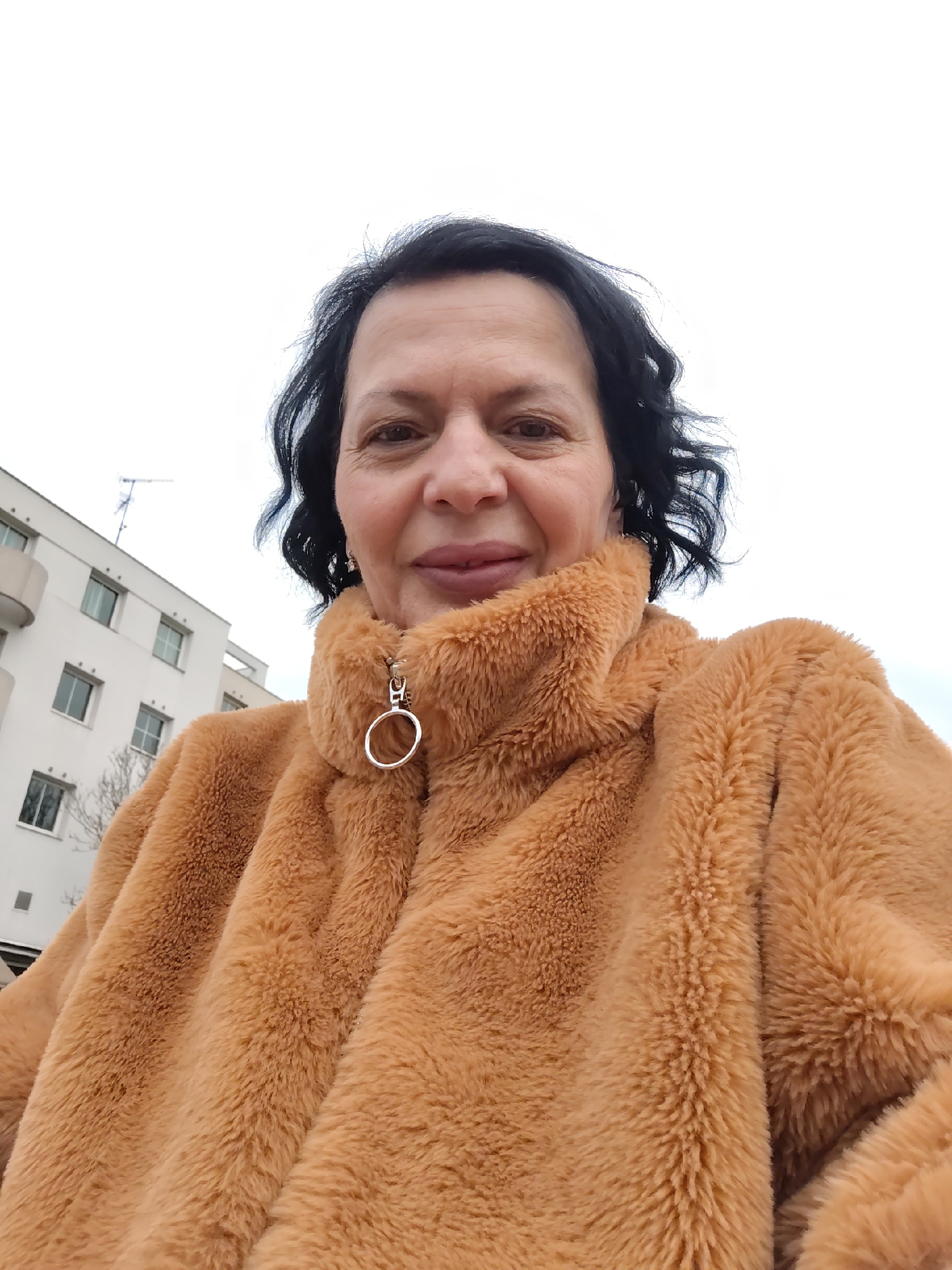 A middle-aged woman with short, dark hair is taking a selfie outdoors. She is wearing a brown, faux-fur jacket with a distinctive circular zipper pull. The background includes a white building with windows and balconies, suggesting an urban residential area. The scene is likely located in Strasbourg, France, based on the building's architectural style and the overall aesthetic. The sky appears overcast.