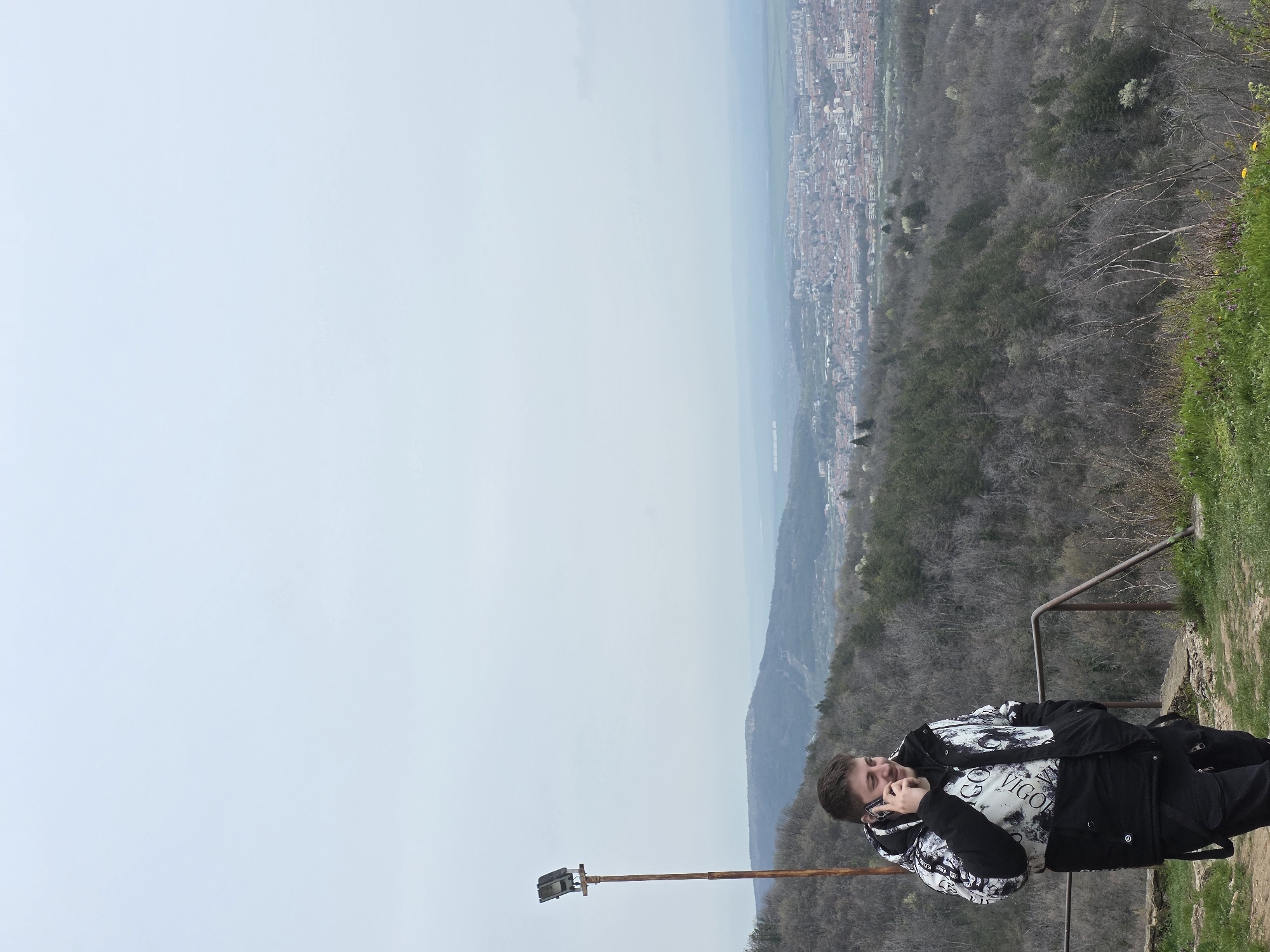 A man is talking on the phone on a scenic overlook in Unknown, Unknown. He is standing on a dirt path with a metal railing, with a view of the city below. The sky is overcast. He is wearing a black jacket with white designs. There is a rusted metal pole with a light fixture next to him. The foreground includes trees and vegetation, and in the distance is a city.
