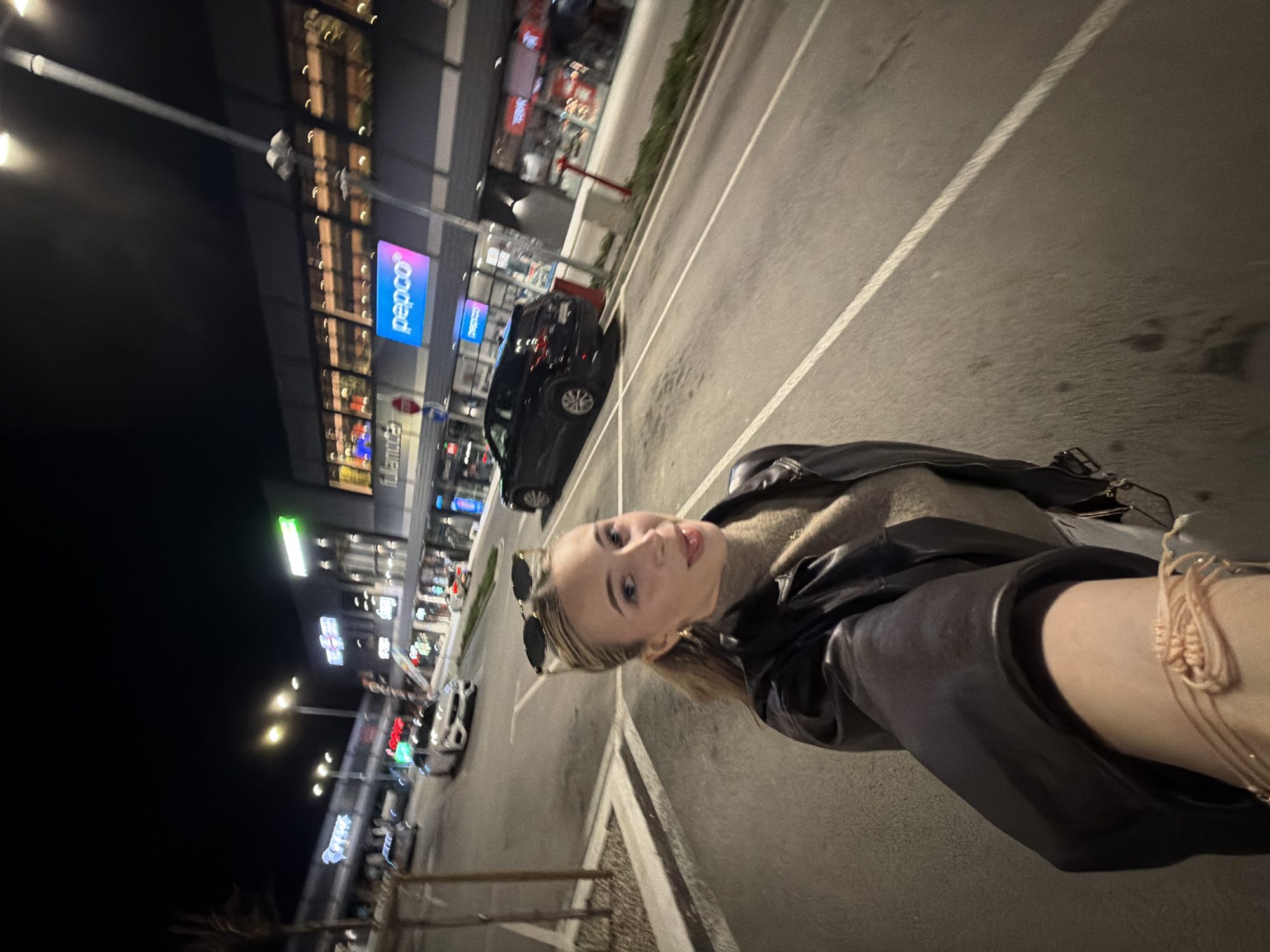 This image captures a night scene at Retail Park Plovdiv in Plovdiv, Bulgaria. In the foreground, a young adult female with blonde hair pulled into a ponytail, wearing a black leather jacket over a light brown turtleneck, is taking a selfie. She has dark sunglasses perched on her head, a light pink braided bracelet on her left wrist, and is looking towards the camera with her tongue slightly extended. The background features a large commercial building with multiple illuminated storefronts, including "fulamoda" and "pepco". An asphalt road with white lane markings and several streetlights separates the subject from the retail complex. Multiple vehicles are visible, including a white sedan in the middle distance and a dark SUV parked further down the road. The sky is dark, indicating nighttime, with artificial light sources illuminating the scene.