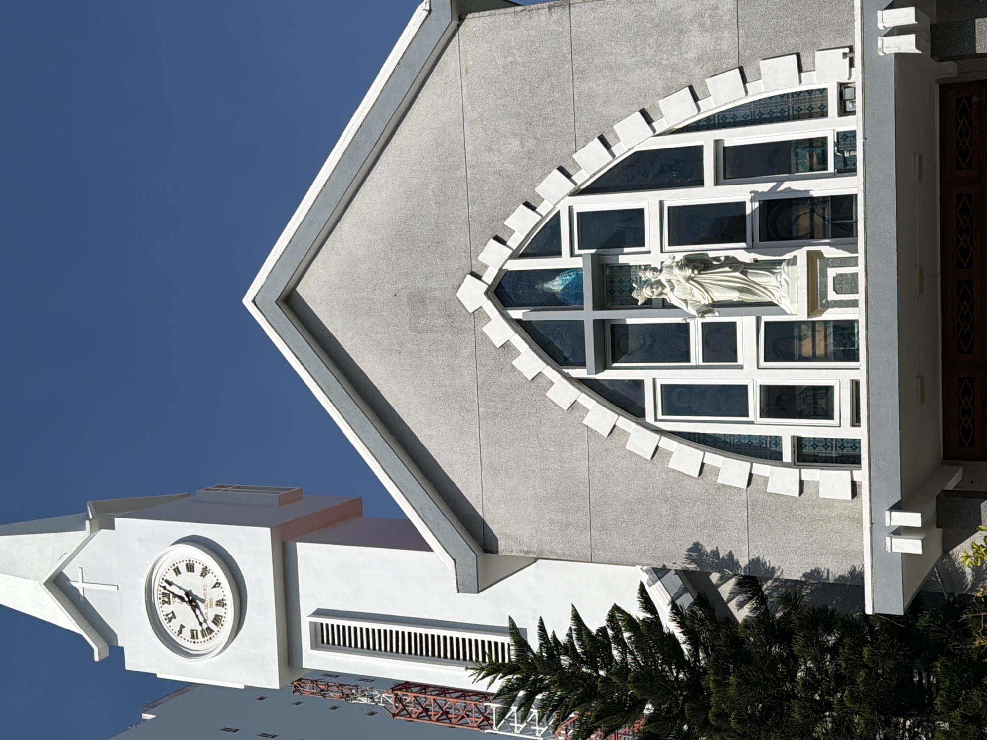 The image shows the facade of a church in Nha Trang, Vietnam. The church has a prominent white clock tower with Roman numerals and hands indicating the time. Below the clock tower is a white statue of Mary holding the infant Jesus, positioned in front of a large, arched stained-glass window. The church itself is constructed of grey textured concrete, with white decorative elements around the arched window. A tall, dark green pine tree stands to the left of the church, partially obscuring a portion of the clock tower and a red and white lattice structure that appears to be a communication tower or scaffolding. The sky is a clear, bright blue.