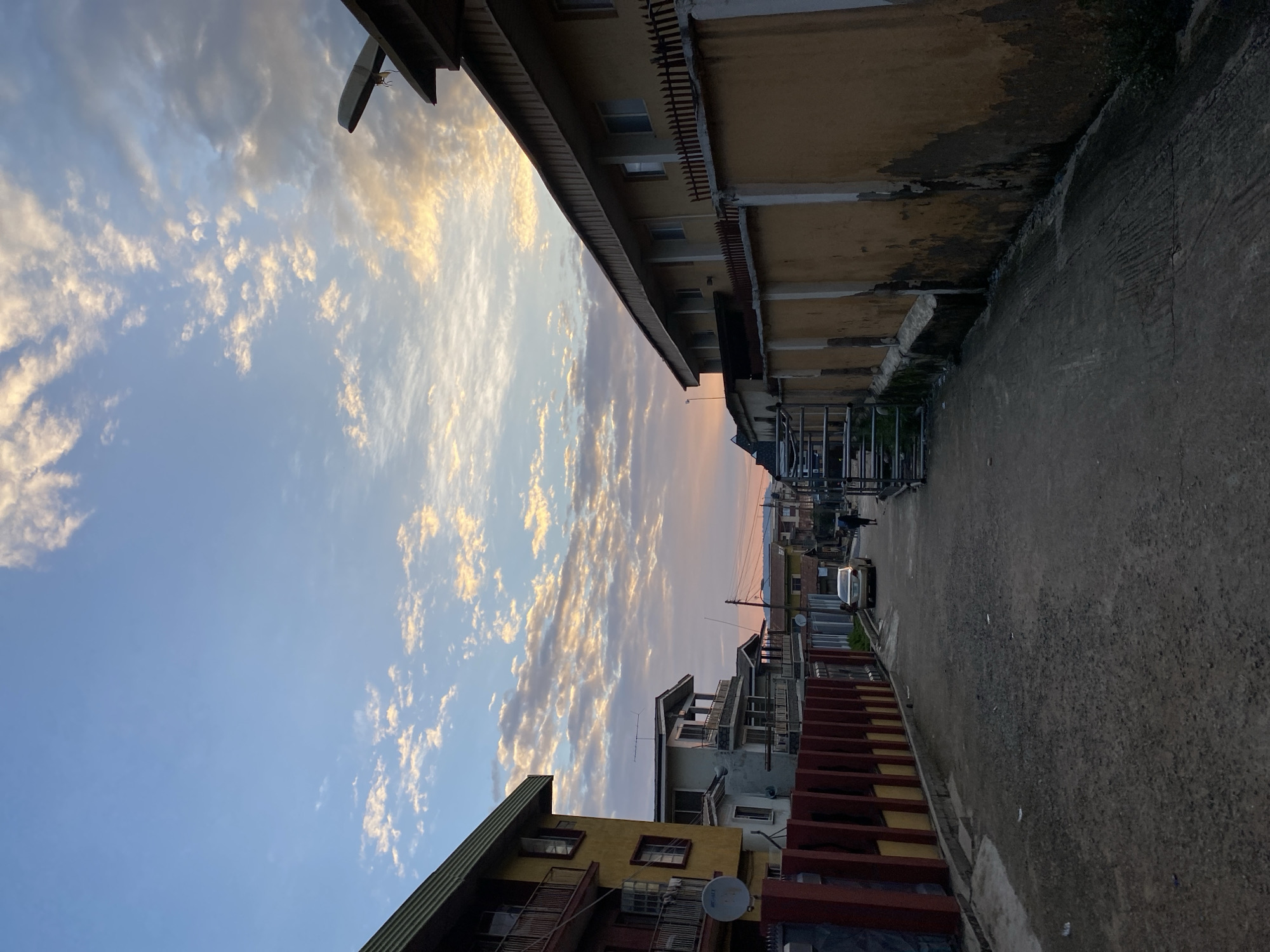 The image depicts a street scene in Abeokuta, Nigeria, during what appears to be either dawn or dusk, indicated by the soft, warm light illuminating the sky and clouds. The perspective is looking down a paved road lined with residential buildings on both sides.
On the left, a yellow building with balconies and windows is prominent, with a red fence in front of it. Further down the street, other buildings of varying heights and architectural styles are visible, some with balconies and satellite dishes.
On the right, a long, yellowish building with a fenced wall runs along the street. The wall shows signs of wear and tear, with patches of discoloration and moss. Metal scaffolding or a structure is visible near the building.
A light-colored car is parked on the road, and a person is walking away from the camera further down the street. The road surface is rough and appears to be a mix of asphalt and gravel. The sky is a mix of blue and scattered clouds, with hints of orange and pink, suggesting the sun is low on the horizon. The overall impression is a glimpse into a residential area in a Nigerian city, capturing the interplay of urban life and the natural beauty of the sky.