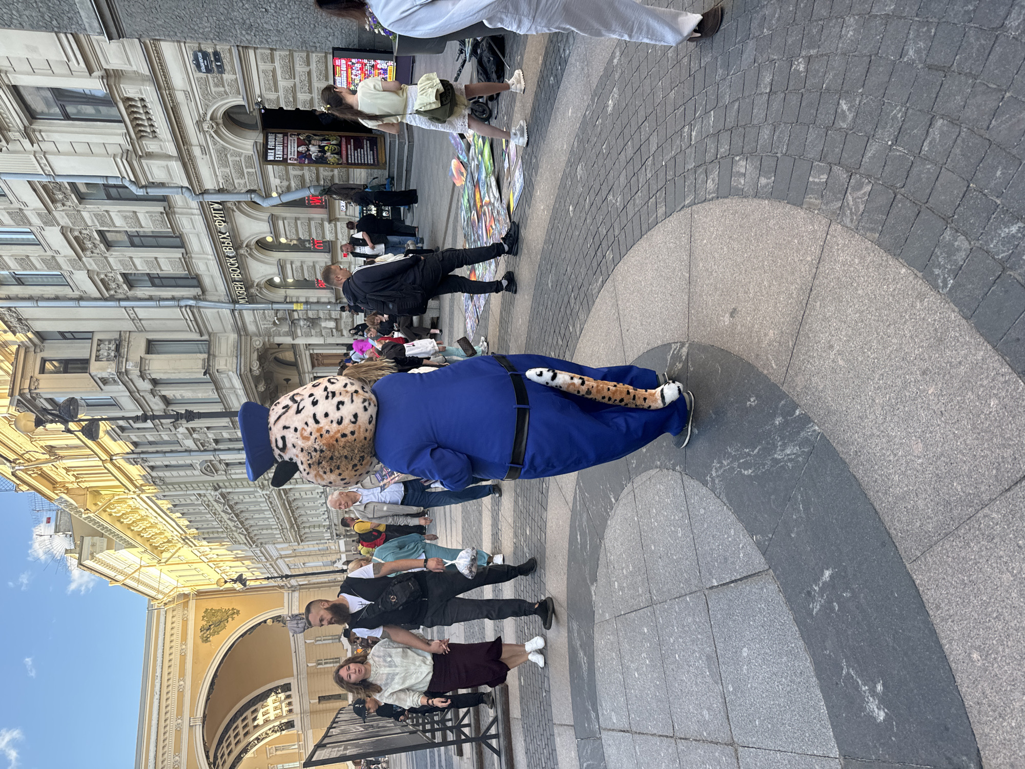 The image depicts a street scene in Saint Petersburg, Russia, likely near Palace Square or Nevsky Prospekt, characterized by the classical architecture and the presence of a large archway visible in the background.
In the foreground, a person in a full-body leopard mascot costume, wearing a blue police-style hat and uniform, is the central subject. The costume features a spotted body, a tail, and a blue jacket with a black belt. The mascot is walking away from the camera.
To the left of the mascot, a couple is walking hand-in-hand. The woman is wearing a light-colored blouse and a dark skirt, while the man is in a dark vest over a white t-shirt and dark pants. They are walking on a patterned stone pavement.
Behind them and further to the left, several other people are visible, including some sitting on stairs. A large display or fence is also present on the far left.
To the right of the mascot, an artist or vendor is displaying various colorful artworks or prints laid out on the ground. A woman in a light yellow top and white skirt stands near the artworks, with a backpack slung over her shoulder. Further to the right, more people are walking, and a sign for a "Wax Figure Exhibition" is visible on a building.
The environment is an urban outdoor setting with a bright sky, suggesting daytime. The ground is paved with different patterns of stone. The buildings in the background are ornate and typical of Saint Petersburg's architecture, with multiple stories and windows.