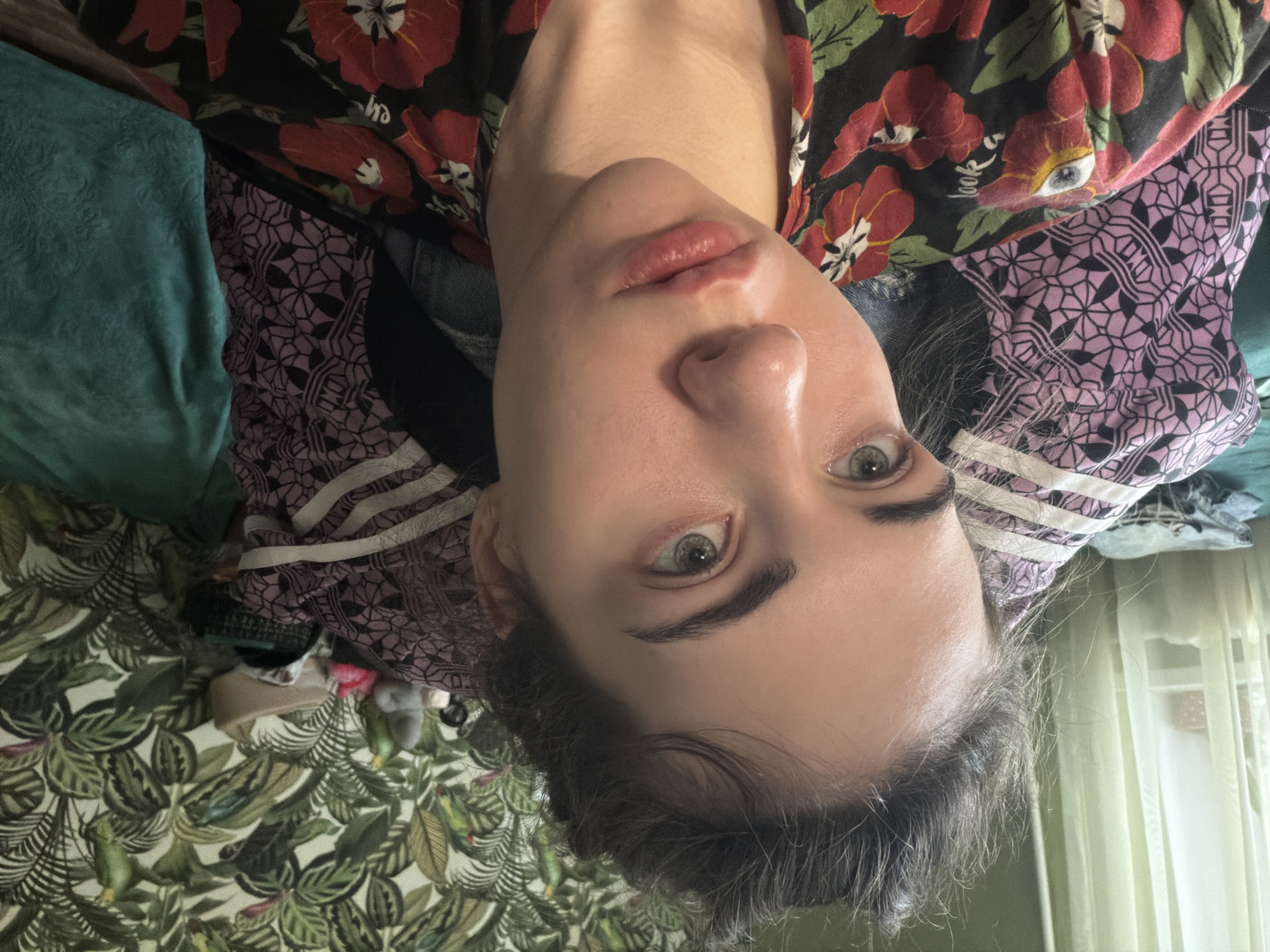 The image is a close-up, selfie-style photograph of a young woman with light skin, light eyes, and dark hair pulled up in a bun. She is wearing a black shirt with a red floral pattern. The background consists of a wall with tropical-themed wallpaper featuring green leaves and parrots, and a pile of colorful blankets and clothing. A window with sheer curtains is visible on the left side of the image. The scene appears to be indoors, likely in a bedroom or living area. There is no identifiable information to suggest the location is in Zavodoukovsk, Russia.
