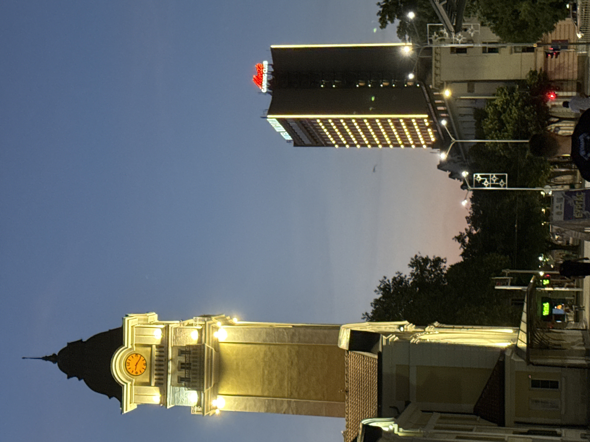 The image captures a twilight scene in the city of Burgas, Bulgaria, specifically on Alexandrovska Street. Dominating the left side of the frame is the historic Clock Tower, illuminated by warm yellow lights, its clock face showing approximately 11:12. Adjacent to it is a building with a tiled roof. To the right, a modern, tall building stands out, its facade outlined with vertical rows of bright yellow lights and topped with a red neon sign that reads "GAMING CLUB". The sky is a deep blue transitioning to a soft pinkish hue near the horizon. Trees line the street, some of which are also illuminated by streetlights. Several pedestrians are visible walking on the sidewalk, some in the foreground and others further down the street. A traffic light is also visible. The overall atmosphere suggests a bustling urban environment in the early evening.