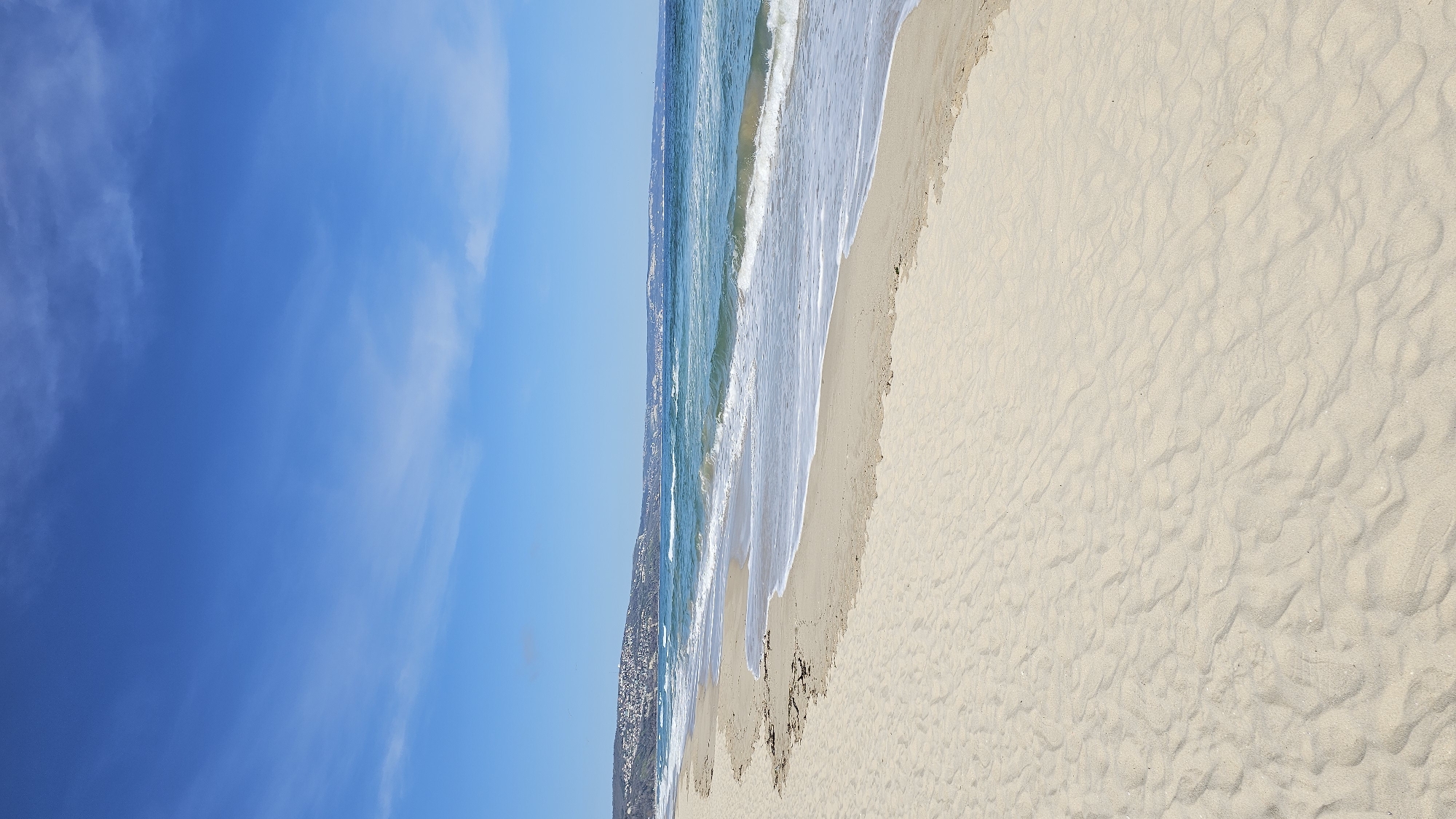 This wide-angle photograph presents a tranquil coastal scene under a clear blue sky. The upper third of the image features a bright, azure sky with delicate, wispy white clouds sparsely spread across it.
Below the sky, the middle section of the image showcases the sea, with waters transitioning from deep blue to vibrant turquoise closer to the shore. White-capped waves can be seen breaking and receding onto the sandy beach. In the distance, a faint, dark, and undulating coastline or landmass is visible, possibly dotted with small, indistinct buildings, suggesting a coastal settlement such as Kranevo, Bulgaria.
The lower portion of the photograph is dominated by the expansive, light beige sand of the beach. The sand's surface displays natural ripples and patterns, along with occasional footprints, indicating the presence of visitors. The overall impression is that of a bright, sunny day, likely during late morning or early afternoon, capturing the serene beauty of the Black Sea coast. No text is visible within the image.