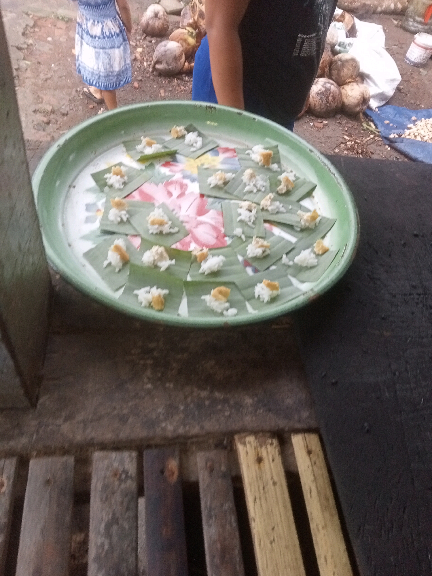 A medium shot captures a large, light green, shallow metal tray filled with small portions of food. The food is presented on individual squares of green banana leaves, each holding a small mound of white rice and a dollop of yellow-orange topping. The tray itself is placed on a dark, textured surface, possibly a table or counter. In the background, the scene is outdoors, with a dirt ground and a few coconuts scattered around. A person wearing a blue and white patterned dress and sandals is partially visible on the left, facing away from the camera. Another person, wearing a dark t-shirt, is also partially visible on the right. The overall impression is a food preparation or serving area in an outdoor, possibly rural, setting.