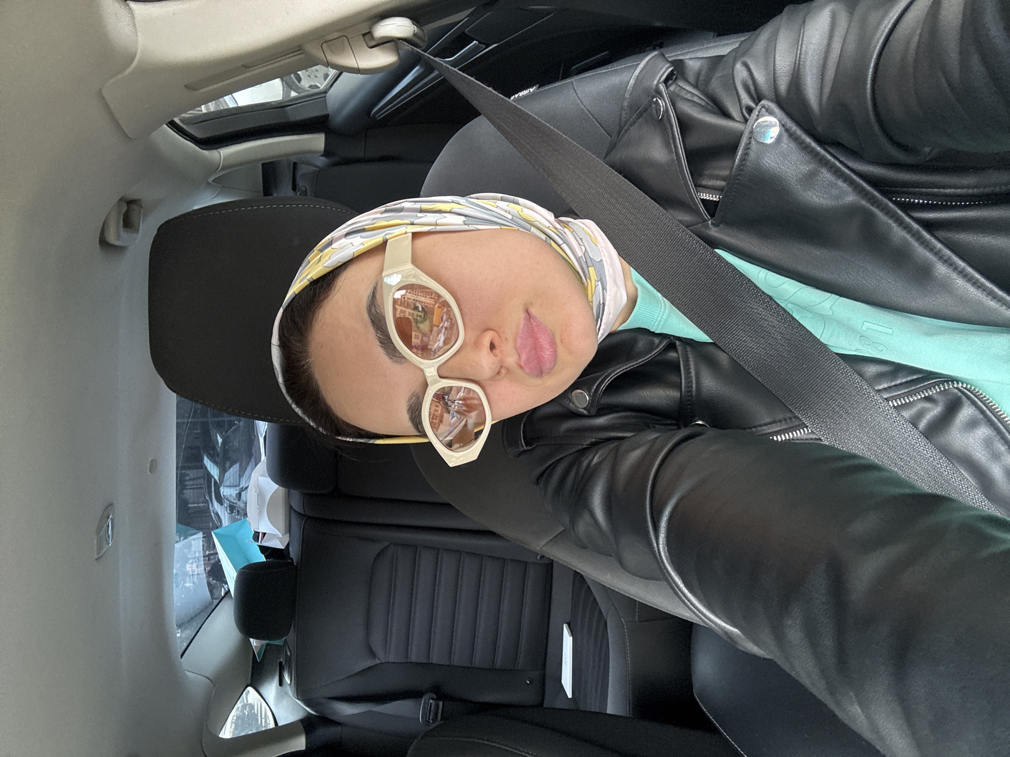 A woman is photographed inside a car. She is wearing a black leather jacket, a turquoise shirt, a headscarf, and sunglasses. A seatbelt is visible across her body. The reflection in her sunglasses suggests the location is Tbilisi, Georgia.
