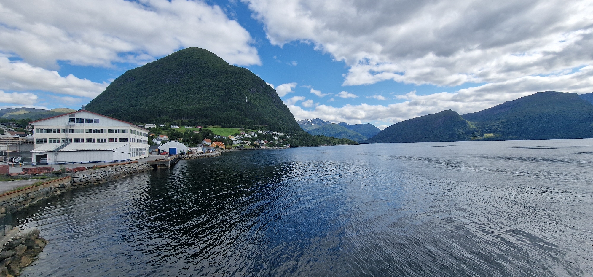 This is a photograph taken from a watercraft, likely a ferry or boat, looking towards the shore of Volda, Norway. The image captures a coastal scene featuring a large body of water in the foreground, reflecting the sky and surrounding landscape. On the left bank, a long, multi-story white building with numerous windows and a red roof is prominent, suggesting it might be a commercial or industrial facility. Adjacent to this building, a rocky shoreline is visible. Further along the coast, a small town or village is nestled at the base of a steep, tree-covered mountain. The town consists of several houses and other structures, painted in various colors, including white, red, and yellow. The mountain rises dramatically behind the town, its slopes densely forested. In the distance, more mountains, some with snow-capped peaks, recede into the background. The sky is partially cloudy, with patches of blue sky interspersed with white clouds. The overall impression is one of a picturesque Norwegian coastal town with a backdrop of imposing natural scenery.
