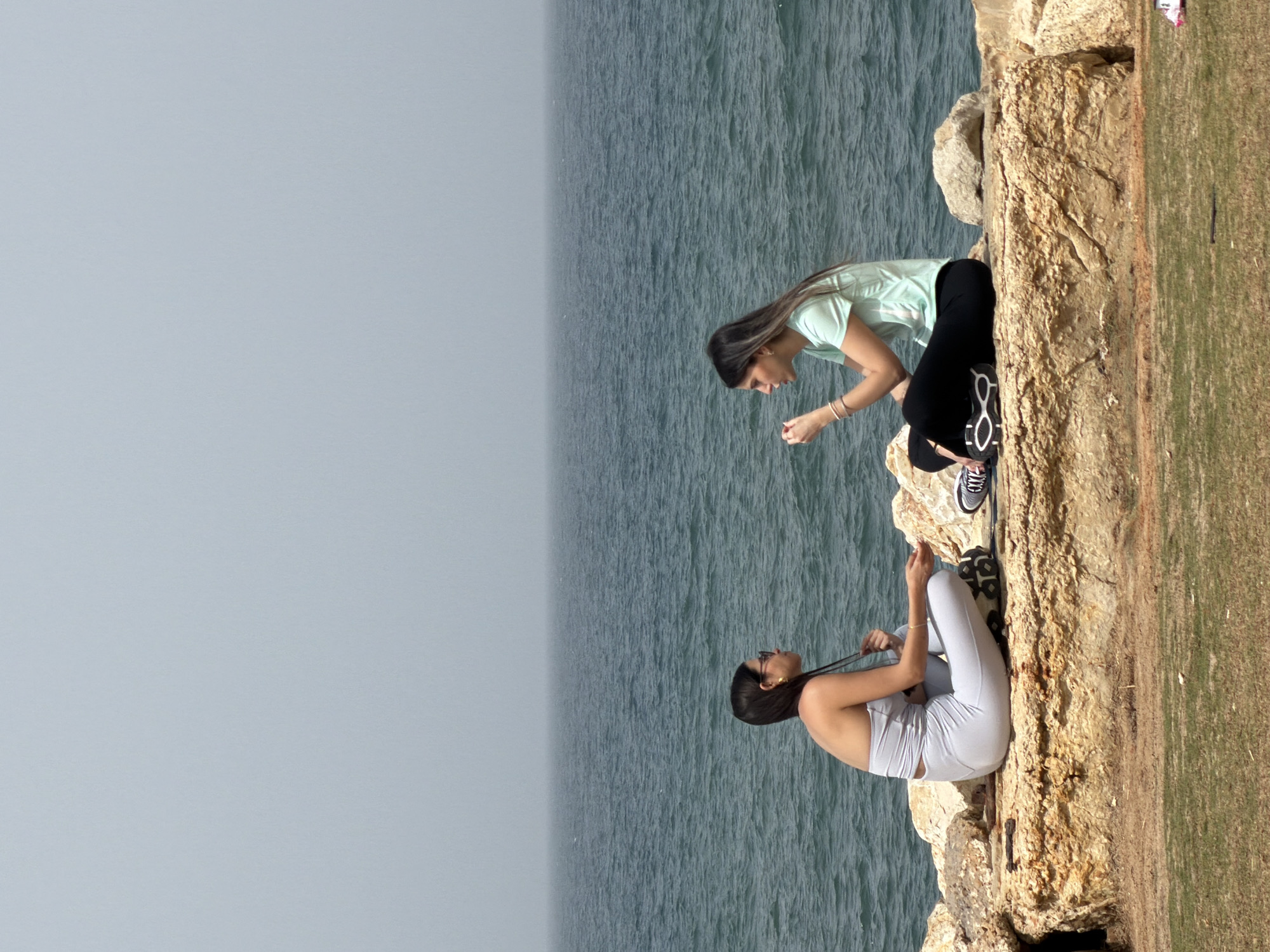 Two women are positioned on a rocky ledge alongside a body of water. One woman is leaning forward, wearing a light green top and black leggings. The other woman is sitting, wearing a light-colored top and matching leggings. They are located in Tel-Aviv, Occupied Palestine.
