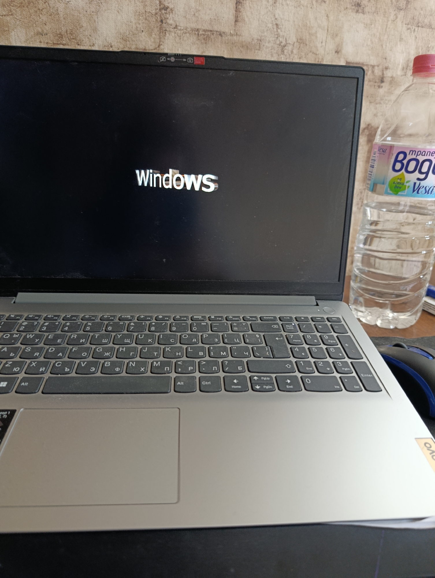 The image shows a laptop computer displaying the Windows logo on a black screen. The laptop appears to be a Lenovo model, indicated by a small sticker visible on the bottom right corner of the base. The keyboard has Cyrillic characters in addition to Latin ones. To the right of the laptop, there is a plastic bottle filled with water; the label on the bottle is partially visible, featuring the word "Vesa" and the word "Bog" in Cyrillic script. A mouse is also visible to the right of the laptop. The background behind the laptop is out of focus and appears to be a textured wall or surface. The image provides no information to determine if this image was taken in the city of Zamfir, Bulgaria.