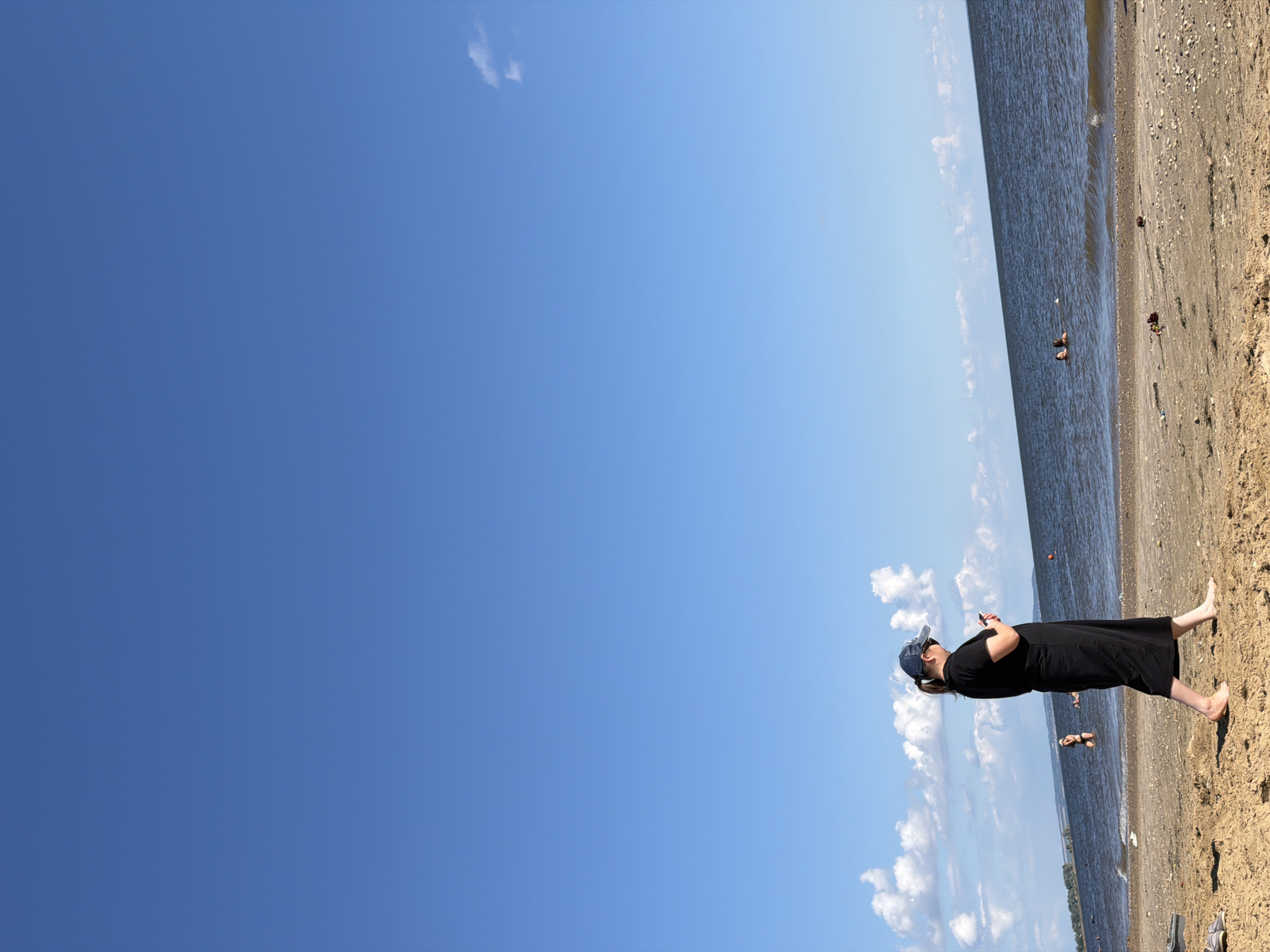 A medium shot shows a woman on a sandy beach, walking and looking at her phone. She wears a black dress and a blue baseball cap. The ocean is to her right, with a few people swimming in it. The sky is clear blue with a few white clouds. The location is Unknown, Unknown.