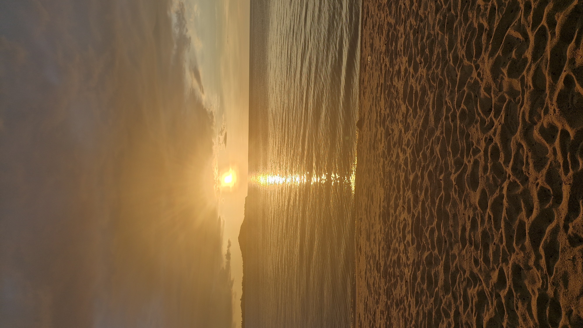 The image shows a serene beach scene at sunset in Baan Bo Phut, Thailand. The sun, a bright orange orb, is low on the horizon, casting a golden glow across the sky and reflecting on the calm water. The sky is filled with wispy clouds tinged with hues of orange and gray. In the distance, a silhouetted island or landmass can be seen. The foreground is dominated by a sandy beach with textured footprints, suggesting recent human activity, though no people are visible in the frame. The gentle waves lap at the shore, creating a peaceful and tranquil atmosphere. The overall impression is one of natural beauty and quietude.