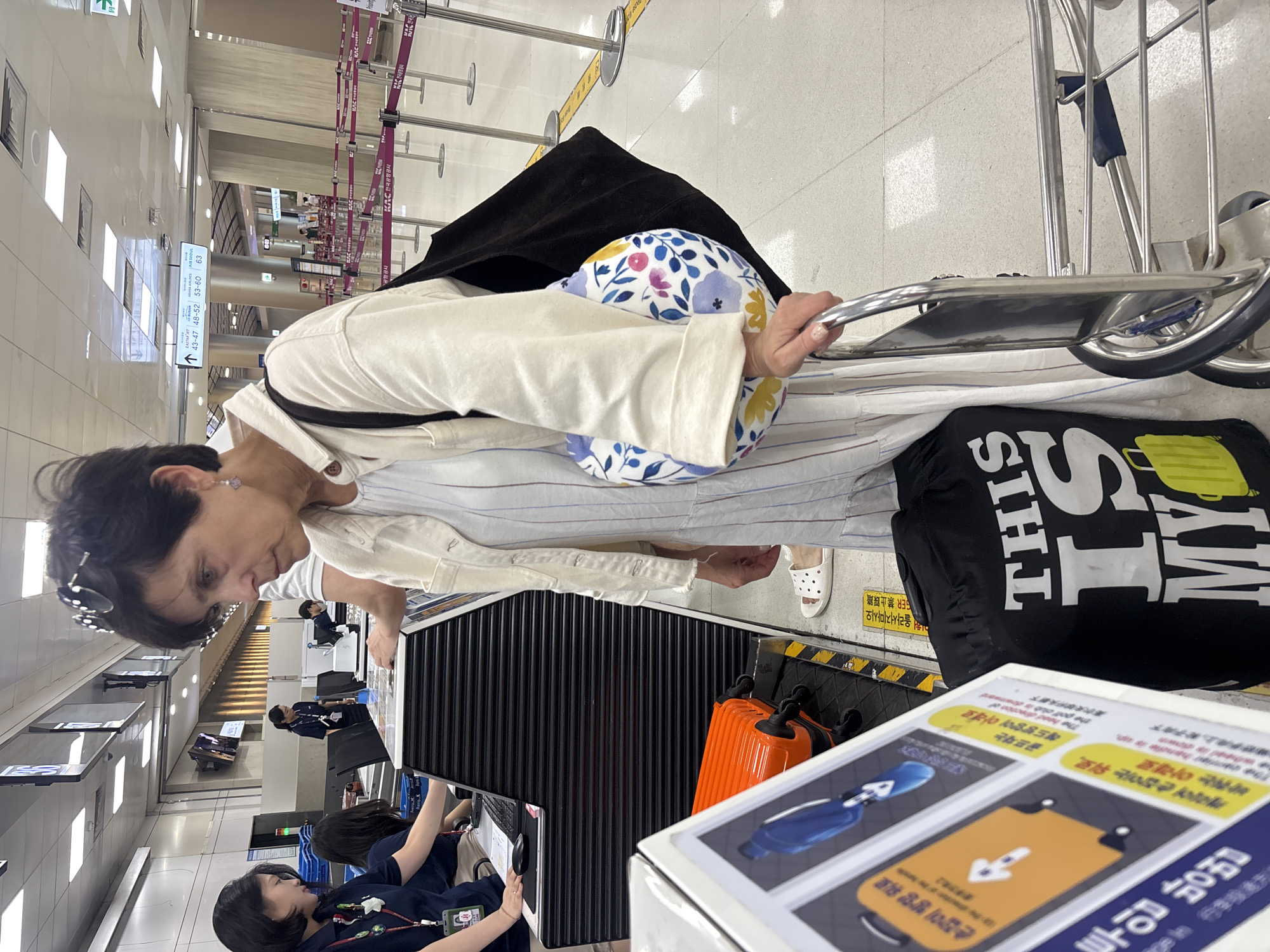 The image shows a woman at a check-in counter in Jeju International Airport, South Korea. She is wearing a light-colored jacket over a striped dress and white slip-on shoes. She is holding onto a luggage cart, and a black bag with white text reading "THIS IS MY" and a yellow suitcase graphic is on the floor next to her. An orange suitcase is on the conveyor belt. Other travelers and airport staff are visible in the background. The environment appears to be a busy airport terminal with check-in desks and queuing lines.
