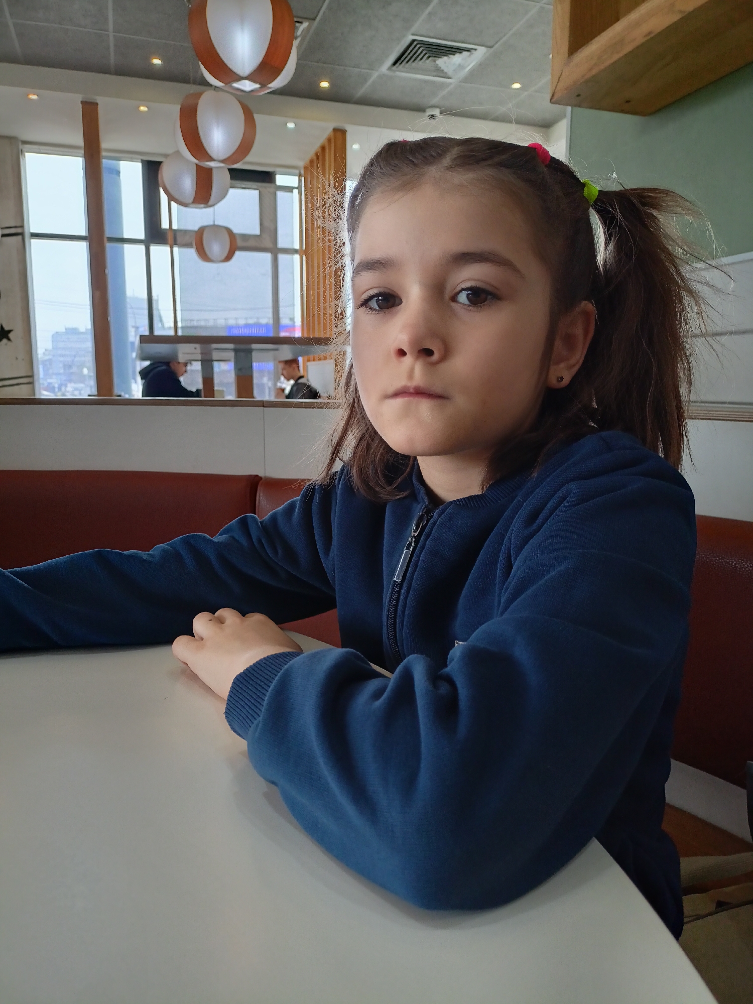 A young girl with brown hair pulled into pigtails is sitting at a white table in an indoor setting. She is wearing a dark blue zip-up sweater. Behind her, there is a red leather bench. Through the windows, there are other people sitting at tables and an urban landscape with buildings can be seen. The interior lighting features decorative hanging lamps with white and brown stripes. The location appears to be a restaurant or cafe.