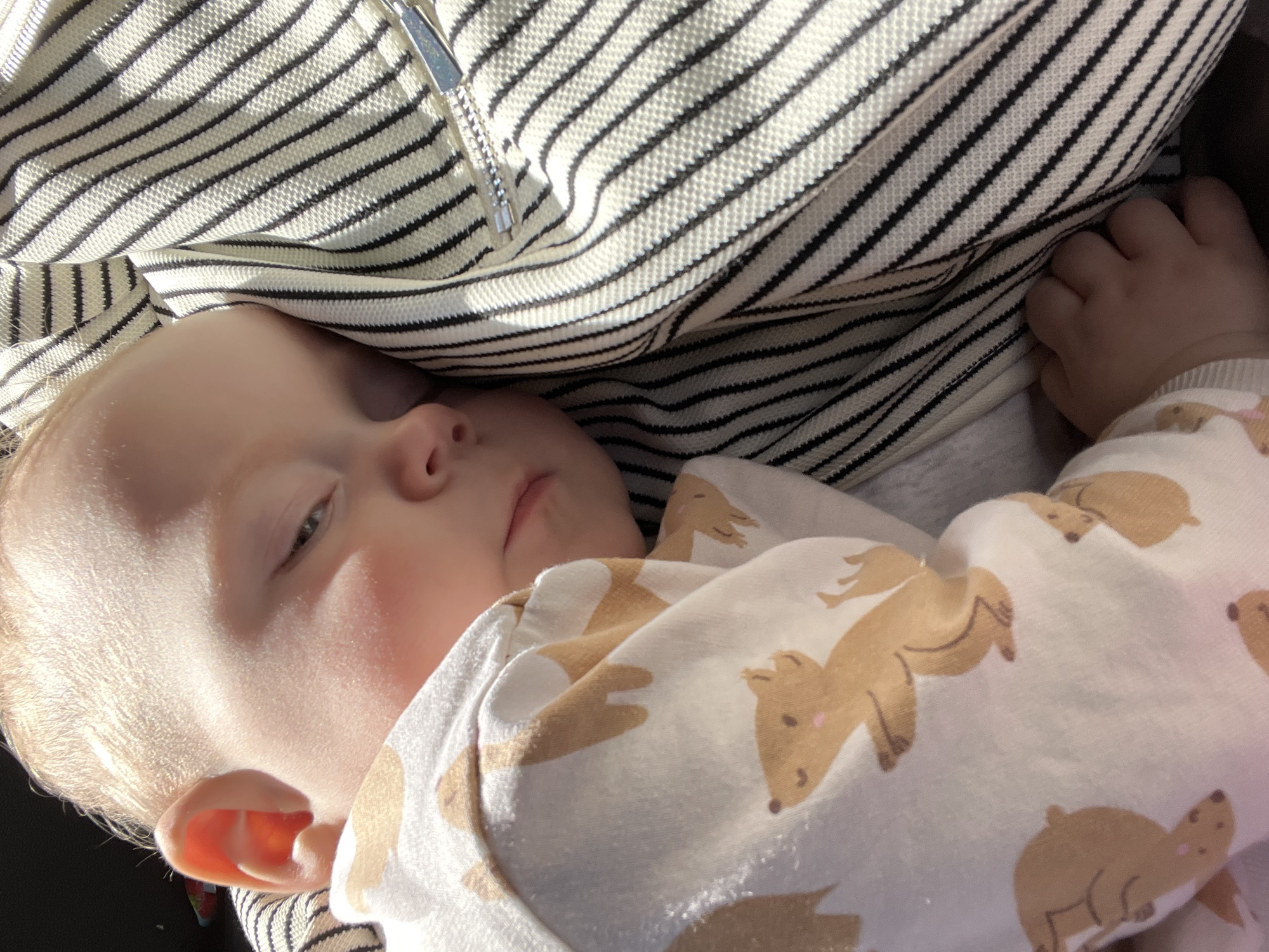 A baby is lying down, partially covered by a striped garment with a zipper. The baby is wearing a white onesie with tan-colored animal illustrations. The baby's right hand is visible, near the striped garment. The location is not identifiable as Perm, Russia, from the visual information.