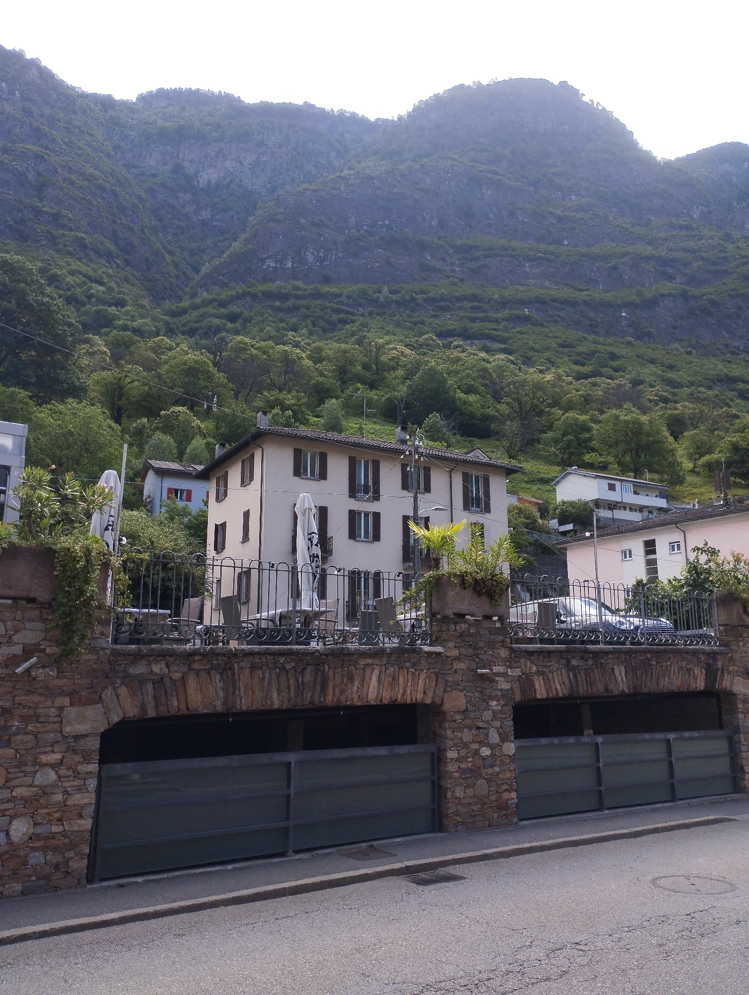 The image depicts a street scene at the base of a steep, tree-covered mountainside. In the foreground, a paved road runs horizontally across the bottom of the frame. To the right of the road, a dark green metal gate spans the width of a garage entrance. To the left of the gate, a stone archway leads into a dark garage. The stone wall supporting the archway and gate is constructed from various sizes and colors of rough-hewn stones.
Above the garage entrance, a decorative wrought-iron fence runs horizontally, enclosing a raised area. Beyond this fence, several buildings are visible. The most prominent is a multi-story building with a pale yellow facade and dark brown shutters on its windows. White umbrellas and outdoor seating are visible on a patio area in front of this building. To the left of this building, another structure with a pale exterior and a red-trimmed window is partially visible. Further to the right, a pink multi-story building stands, and behind it, a light blue and white building with multiple balconies is situated on the hillside.
The upper portion of the image is dominated by the lush green slopes of a mountain, densely covered with trees. The sky above is bright and slightly hazy, suggesting it is daytime, possibly late morning or early afternoon. No people are visible in the image.