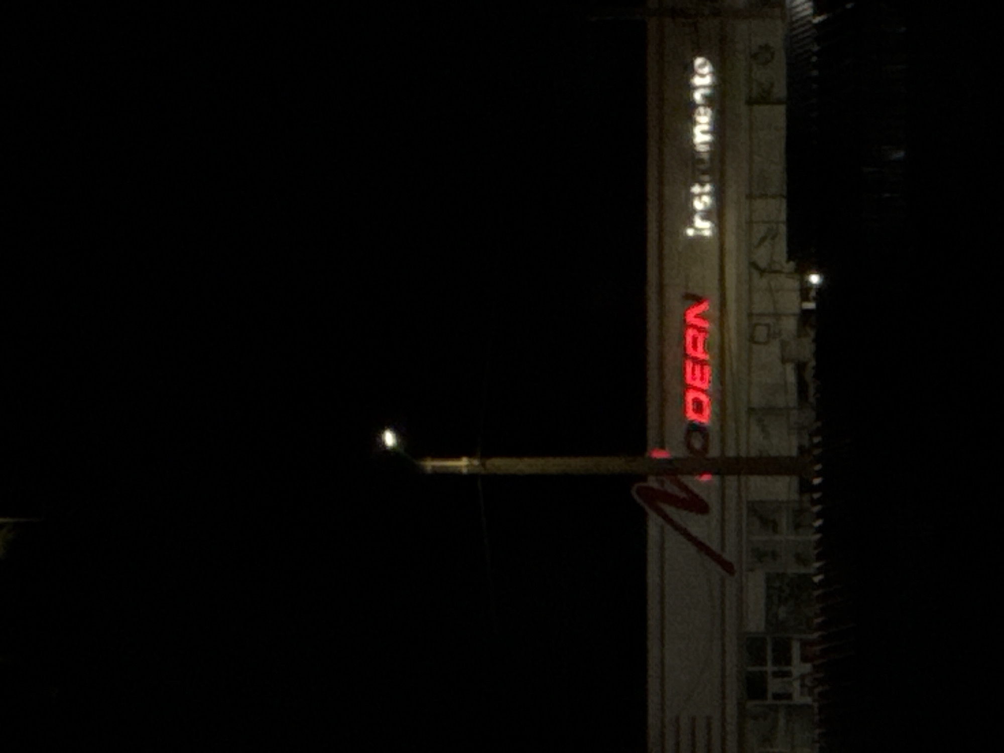 The image shows a building at night with illuminated signage. The building appears to be a commercial establishment, possibly a store or office, with large windows and a horizontally oriented facade. A streetlamp with a bright light is visible on the left side of the image, casting illumination on the scene. Above the windows, a sign in neon letters reads "Modern instrumento". The "Modern" part is in red, and "instrumento" is in white. A dark, shadowy fence or barrier runs across the foreground of the image, obscuring the lower portion of the building. The overall scene is dark, characteristic of nighttime, with the only significant illumination coming from the streetlamp and the building's signage. Based on the signage, the location is likely a business named "Modern Instrumento" in the city of Cahul, Moldova.