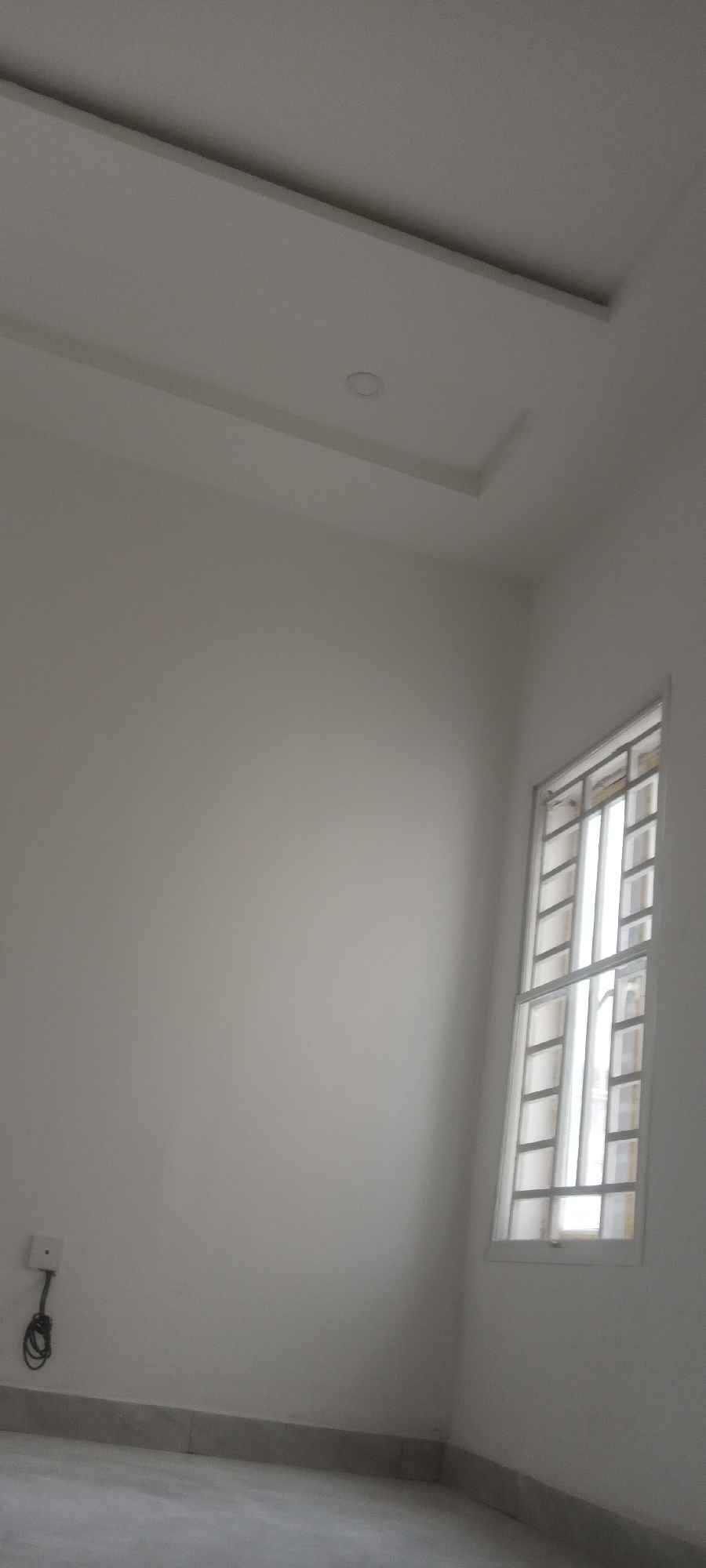 A room in Unknown, Unknown, features a white wall, a window with bars, and a power outlet with a cable plugged in. The ceiling is white with recessed lighting and a rectangular design element. The floor is gray.