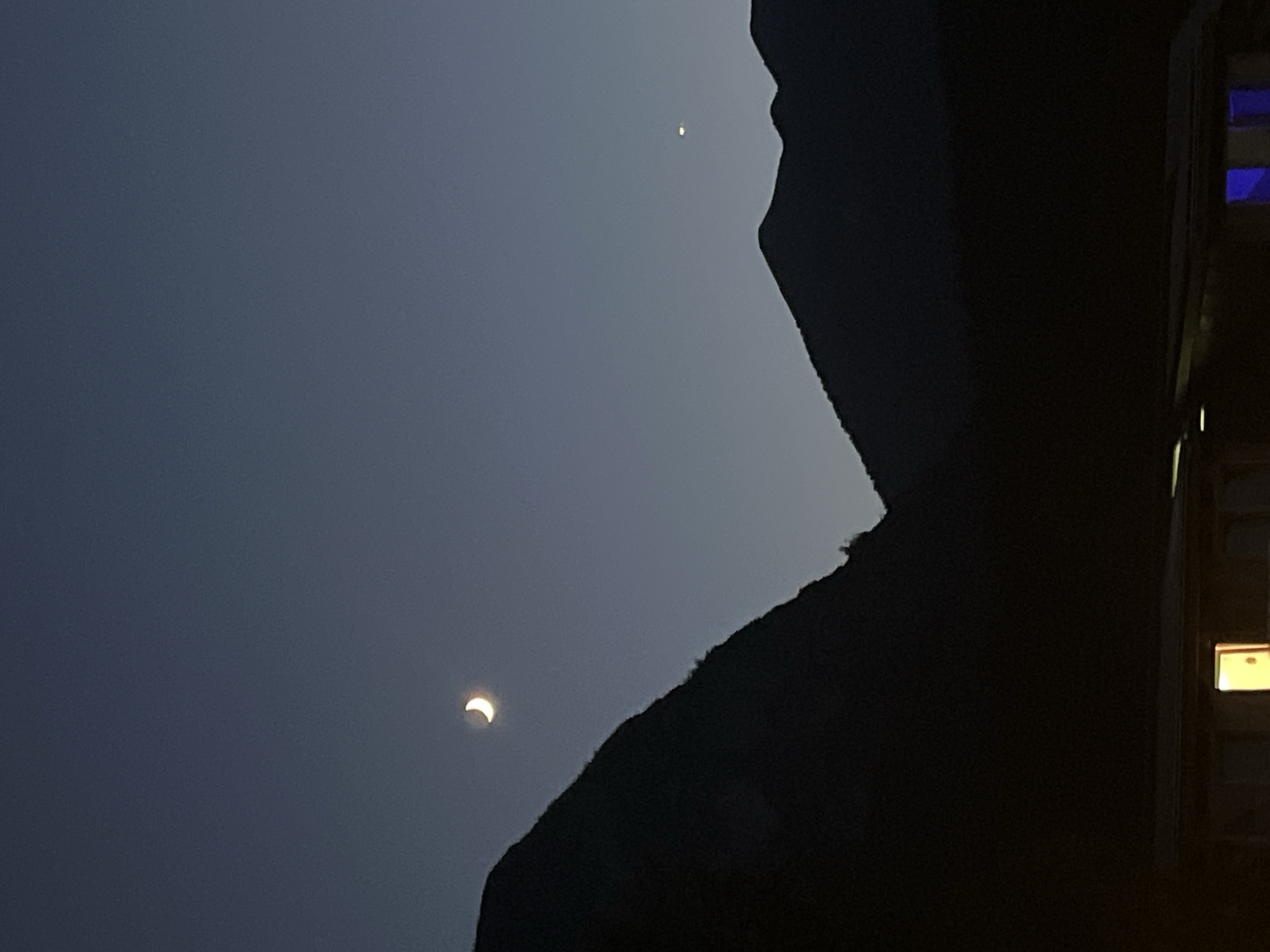 This image captures a twilight scene in Garmisch-Partenkirchen, Germany. Silhouetted against a gradient of darkening blue sky are the dark, rugged outlines of mountains. A crescent moon is visible in the upper left quadrant of the sky, emitting a soft, pale yellow light. Further to the right, a small, bright point of light, likely a planet, is visible. In the foreground, the dark silhouette of a building or structure is present. A single window on this structure emits a bright yellow light, indicating an interior light source. To the right of this illuminated window, another window glows with a vibrant blue light. The overall impression is of a serene, natural landscape at the cusp of night, with signs of human habitation in the foreground.
