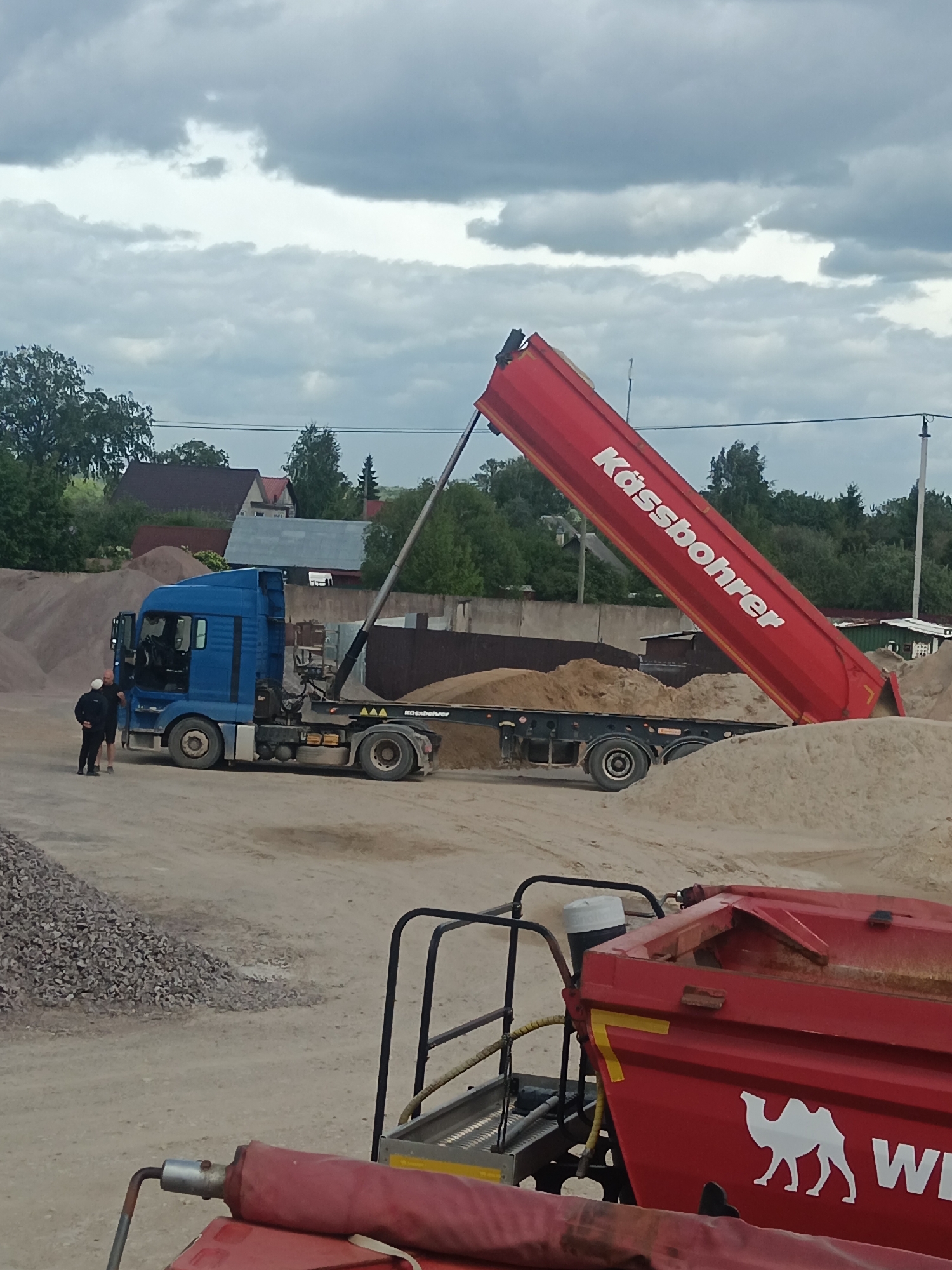 The image shows a blue MAN truck with a red Kässbohrer tipper semi-trailer in Яльгелево, Russia. The tipper is raised and is unloading sand. Two men are standing near the truck cab, engaged in conversation. In the foreground, there is a portion of another red trailer with a white camel logo. Gravel is piled to the left of the truck and sand piles surround the trailer. In the background are buildings and trees. The sky is overcast.