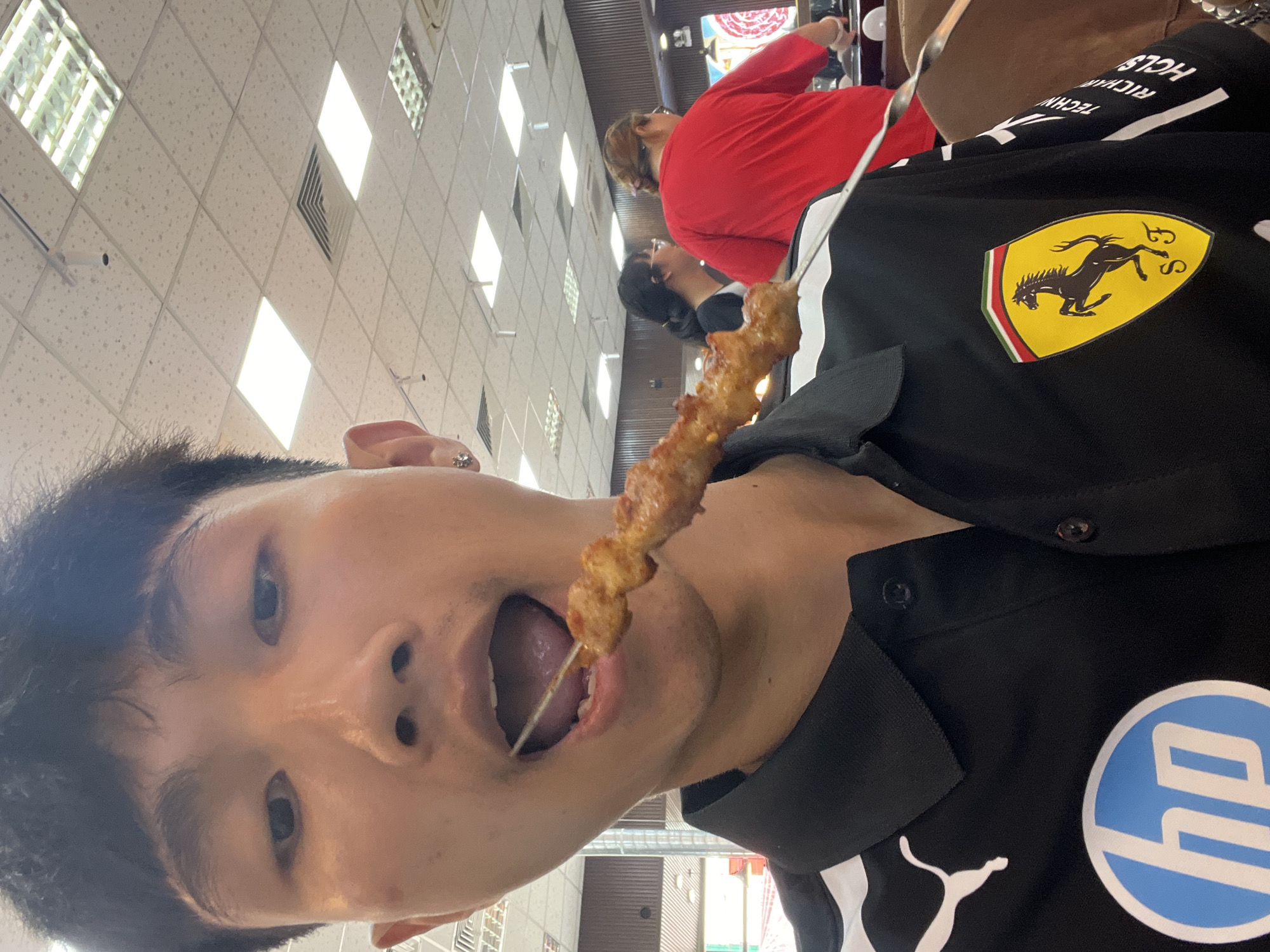 A person is holding and eating a meat skewer indoors. The person is wearing a black shirt with a Ferrari logo on the chest, an HP logo, and a Puma logo. Other people are in the background. The location appears to be an indoor restaurant. Based on visual cues, the location is suspected to be in Đà Nẵng, Vietnam.