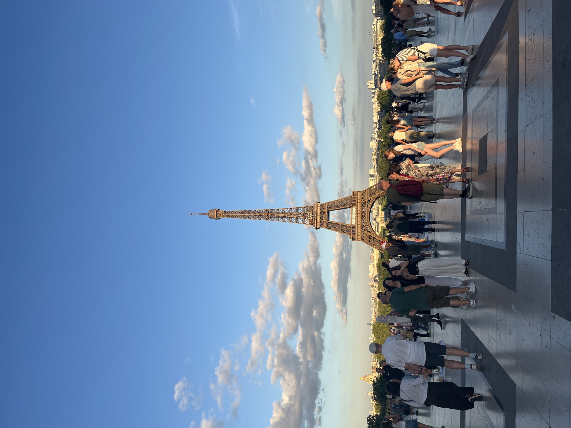 A photograph taken in Paris, France, shows a crowd of people gathered near the Eiffel Tower. The image is oriented vertically, with the Eiffel Tower prominently featured on the left side. The sky is a clear blue with scattered white clouds. The Eiffel Tower is a metallic lattice structure, with its intricate design visible.
In the foreground, a diverse group of people are standing and walking on a paved area. They are dressed in casual summer clothing, suggesting a warm day. Some individuals are posing for photos, while others are engaged in conversation or simply observing the surroundings. The people are of various ages and ethnicities, all contributing to a lively atmosphere.
The background reveals the cityscape of Paris, with buildings and trees lining the horizon. The sunlight casts long shadows, indicating that it is either early morning or late afternoon. The mood of the scene appears relaxed and joyful, with many people smiling and enjoying their time.
The pavement is made of large stone tiles arranged in a geometric pattern, adding to the urban aesthetic. The overall impression is of a popular tourist destination during a pleasant day, where people from all over the world come to experience the iconic landmark. No visible text is present in the image.