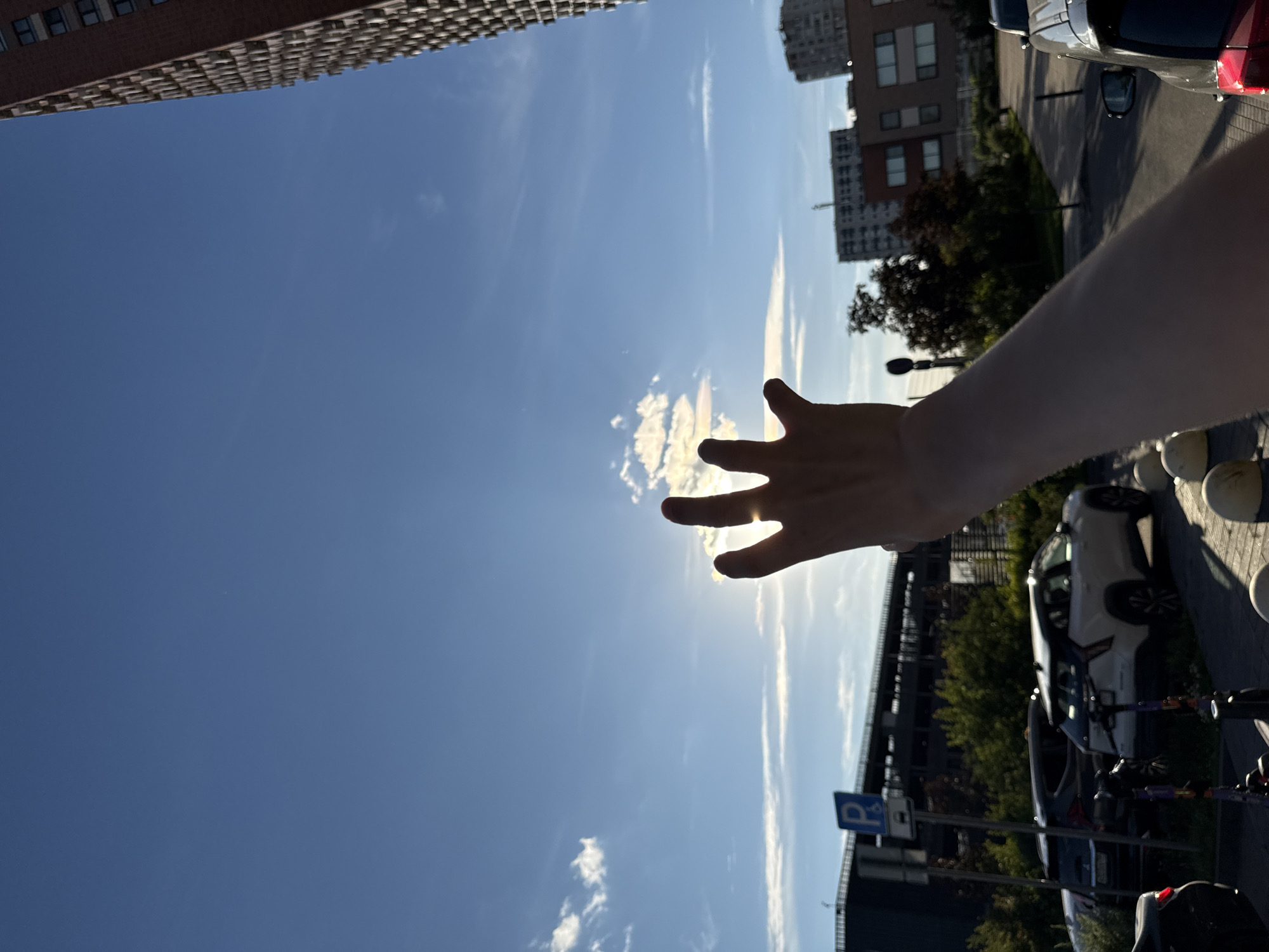 In Moscow, Russia, on a clear day with scattered clouds, a person's right arm and hand are raised towards the sky, obscuring a portion of the sun which is partially visible behind their fingers. The sky is a vibrant blue, with a soft, iridescent halo forming around the sun, suggesting a cloud iridescence phenomenon. In the background, a tall, multi-story brick building with numerous windows stands on the right side. To the left, a parking lot is visible with several cars parked, including a white SUV and a dark SUV. A blue parking sign with a white "P" is also present. Trees and other buildings are discernible further in the background. The overall scene captures a moment of reaching out towards the sun amidst an urban environment.