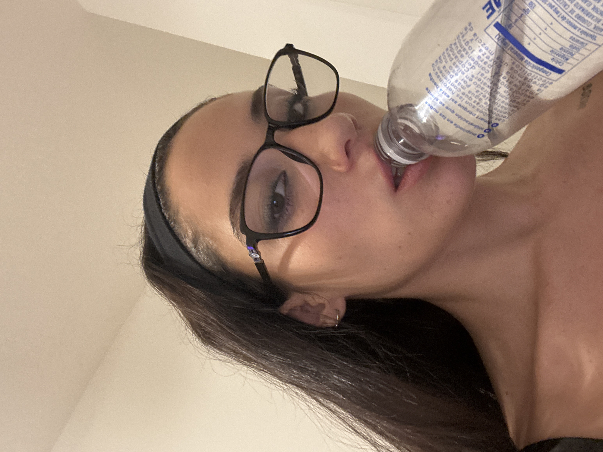 A woman with long brown hair, wearing glasses and a black headband, is drinking from a plastic water bottle. The bottle label has some text in Spanish, suggesting the location could be in a Spanish-speaking region. The woman is wearing a dark-colored shirt and has a light skin tone. The background is a plain, light-colored wall. It's not possible to determine the exact location in Buenos Aires from this image.