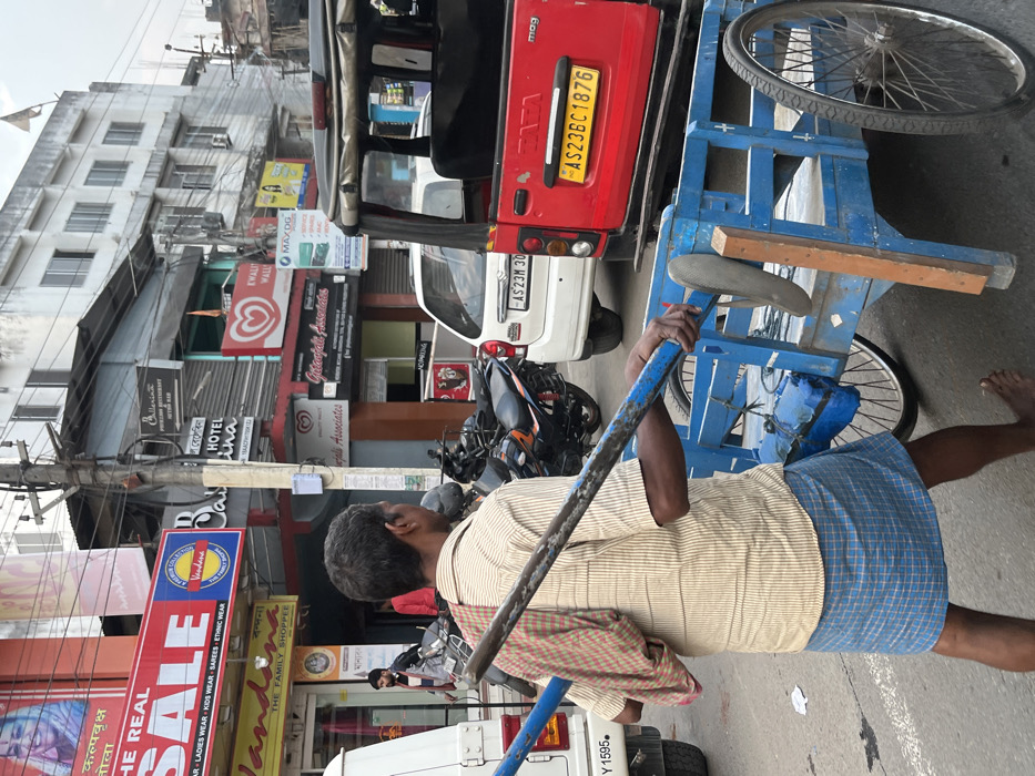 This image, set on a busy street in Tinsukia, India, captures a vibrant urban scene during daylight hours. The primary subject is a man, viewed from behind, wearing a striped light-colored shirt and a blue and white checkered lungi (or similar wrap). He is barefoot and holds a long blue metal pole, which appears to be part of a large, blue, two-wheeled handcart or goods carrier that he is pulling or pushing. A red and white checkered cloth is tucked into his shoulder.
The street is bustling with vehicles and commercial activity. Immediately to the left of the man, a red auto-rickshaw is parked or moving, displaying the license plate "AS23BC1876," indicating registration in Assam. A white car with a partial license plate "AS 23M 30" is visible behind the auto-rickshaw, and a black motorcycle is parked further down.
Numerous multi-story buildings line the street, featuring a variety of shops and businesses, adorned with colorful signs. Notable visible text from these signs includes "THE REAL SALE" for "LADIES WEAR KIDS WEAR SAREES ETHNIC WEAR," "Jandana THE FAMILY SHOPPEE," "HOTEL Lina," "Ballerina," and "MAXDG." An "♡ Kwality Wall's" ice cream sign is also prominent. Other texts like "कल्पवृक्ष" (Kalpavriksha) and "तोला" (Tola) are also visible. "Vefali Associates" and "NOPRING" (likely "No Parking") are also present. Utility poles with a dense network of overhead wires run along the street, a common sight in Indian cities. The overall impression is one of a functional, active commercial thoroughfare.