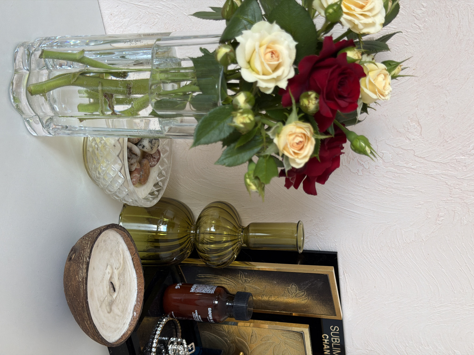 A still life arrangement is displayed on a white surface against a textured, pale pink wall. In the foreground, a clear glass vase holds a bouquet of red and cream-colored roses with green leaves and buds. To the right of the vase, a decorative clear glass bowl contains small stones. Further to the right, a green glass decorative object with a stacked, rounded shape stands next to a small brown bottle with a black cap. Behind these items, a black framed picture with gold detailing is visible, along with another gold-framed picture and a dark frame with text that appears to read "SUBLIM..." and "CHAN...". In the immediate foreground on the right, a lit candle made in a coconut shell sits on the white surface. The overall scene suggests an indoor decorative setting. The location of Rossiyskiy, Russia is not depicted in the image.