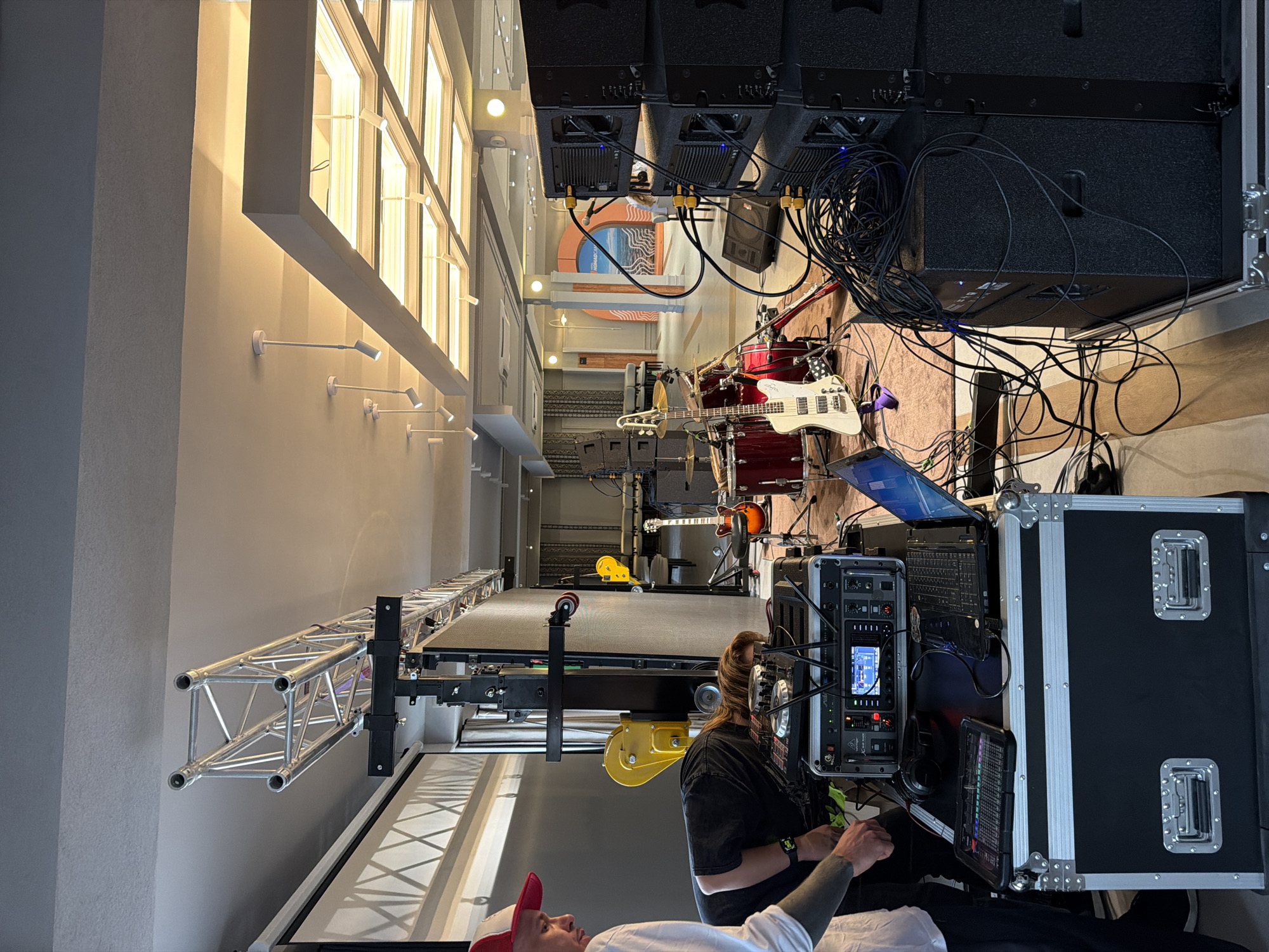 The image depicts an indoor scene, possibly a venue or event space, with several individuals setting up audio equipment. In the foreground, two individuals are visible. One, wearing a white shirt and a red and white cap, stands to the left, looking down. Another person, wearing a dark t-shirt, is bent over a sound mixing console and other electronic equipment, appearing to adjust settings. Various audio components, including speakers, a laptop, a mixing board, and headphones, are arranged on a black flight case.
In the background, musical instruments such as a drum set and guitars are set up, suggesting preparation for a musical performance. Further in the background, the space appears to be a large room with lighting fixtures on the ceiling, furniture, and decorative elements. The overall impression is of a setup in progress for an event. The location in Каджи-Сай 2, Kyrgyzstan, is not visually evident from the image itself, as no identifiable landmarks or textual cues related to the specific city are present.