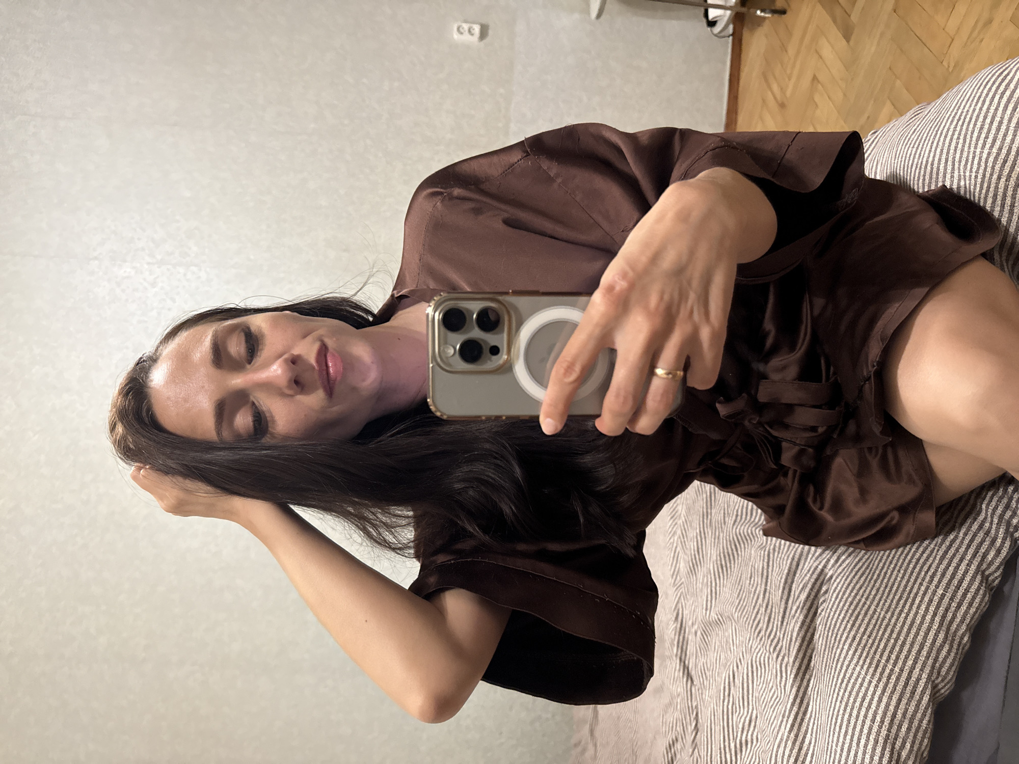 A woman is taking a selfie in a bedroom, holding a smartphone in her right hand. She is sitting on a bed with striped bedding. The woman has long, dark hair and is wearing a brown satin robe. Her left hand is raised and touching her hair. The background shows a wall with textured wallpaper and a power outlet. The floor is wooden parquet. The scene suggests an indoor setting in a private residence. There is no discernible information to place the location in Saint Petersburg, Russia.