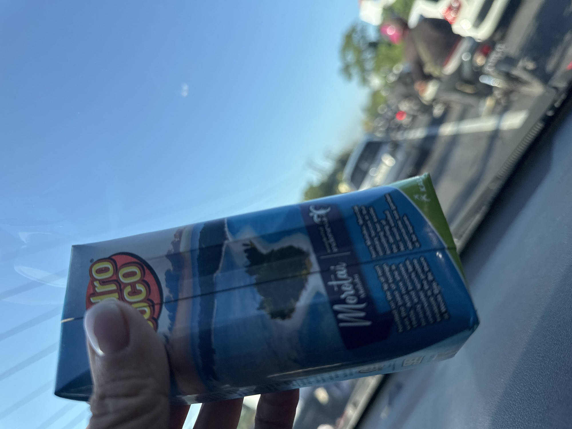 The image shows a hand holding a juice box with the brand name "Hydro Coco" and the word "Morotai" on it. The background is blurred, showing what appears to be traffic on a road, possibly viewed through a car window. The environment suggests an outdoor setting with clear sky. There's no explicit information to ascertain the location as the city of Denpasar, Indonesia.