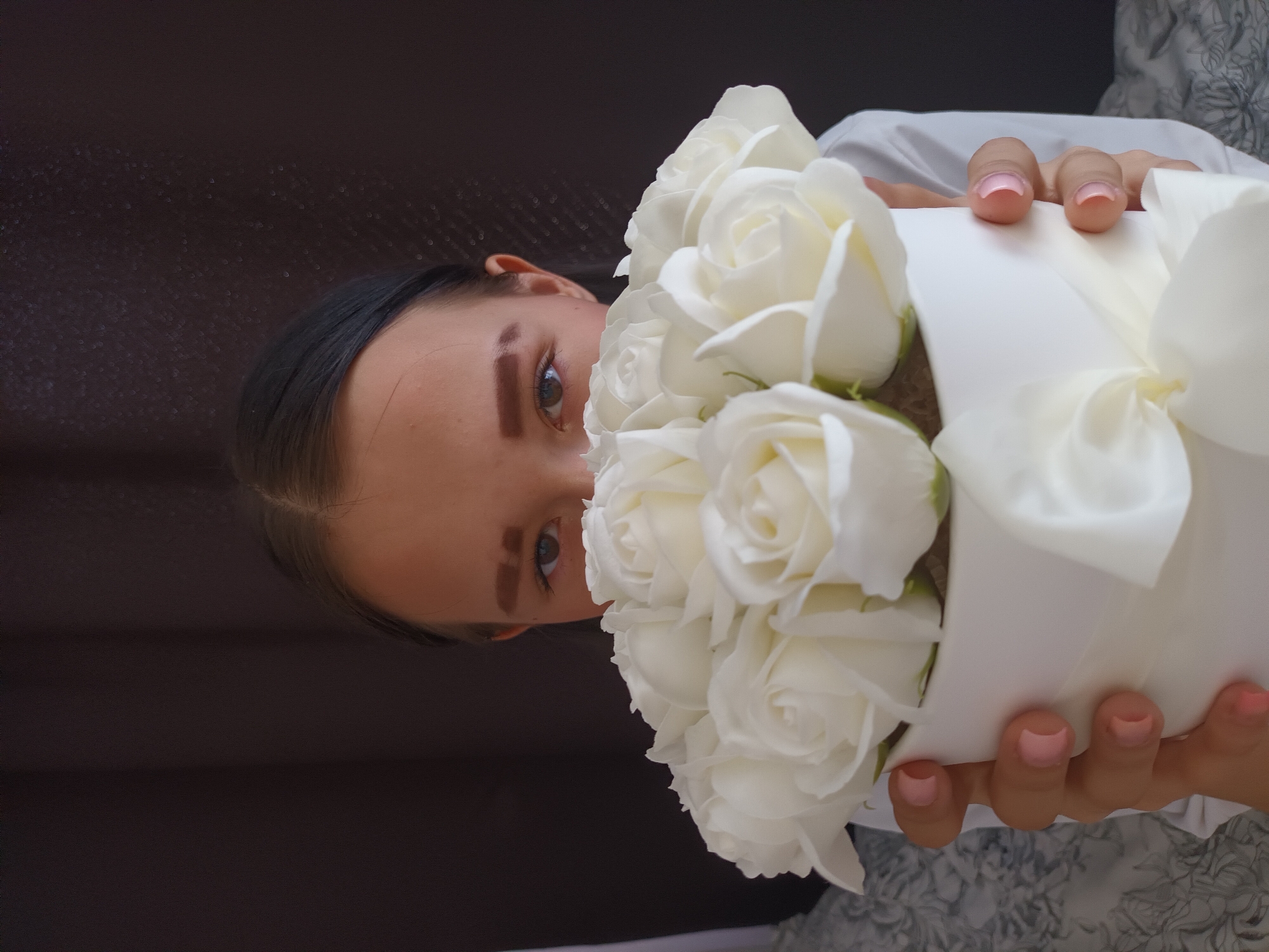A medium, close-up shot depicts a woman holding a box of white roses. Only the woman's forehead, eyes, and part of her left ear are visible. She has brown eyes and her dark hair is pulled back. The woman's hands, with pale pink nail polish, hold a white, round box filled with white roses. A large, white bow adorns the box. The background consists of a dark brown curtain. The location is Ольховка, Russia.