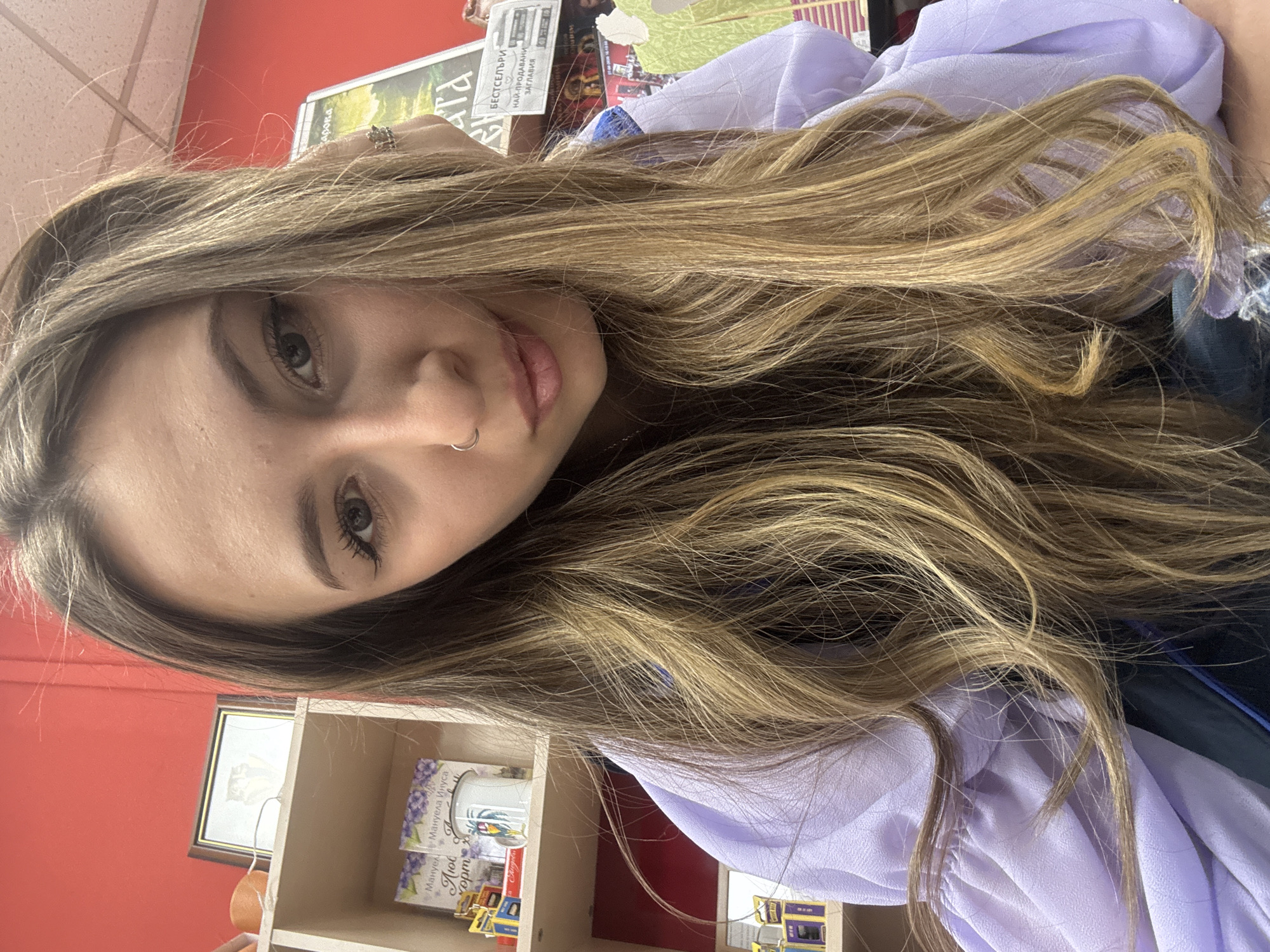 A close-up, selfie-style portrait features a young person with long, wavy, light brown and blonde hair, rotated 90 degrees to the left. The subject is a female with fair skin, dark eyes looking directly at the camera, and light pink lips slightly parted. She has a silver ring piercing her left nostril. Her left hand is partially visible near her hair, wearing a metallic ring on one finger. She is dressed in a light purple, possibly ruffled, top.
The background suggests an indoor setting, likely a bookstore or a room with shelves. A prominent red wall takes up much of the background space, with a small framed picture hanging on it. To the right, a light-colored wooden shelving unit holds several books and a white mug with a colorful parrot-like design. Visible text on one book or magazine cover reads "БЕСТСЕЛЪРИ НАЙ-ПРОДАВАНИ ЗАГЛАВИЯ" (Bestsellers - Top Selling Titles) in Cyrillic. Another book cover partially displays "Мануела Иисуса" (Manuela Jesusa) and "Любов" (Love). The Cyrillic text is a strong cultural cue, consistent with the given location context of Veliko Tarnovo, Bulgaria.
The activity depicted is the subject posing for a photograph, likely a self-portrait. The lighting is bright and even, illuminating the subject's face. The overall mood is casual, with the person displaying a neutral to slightly soft expression.