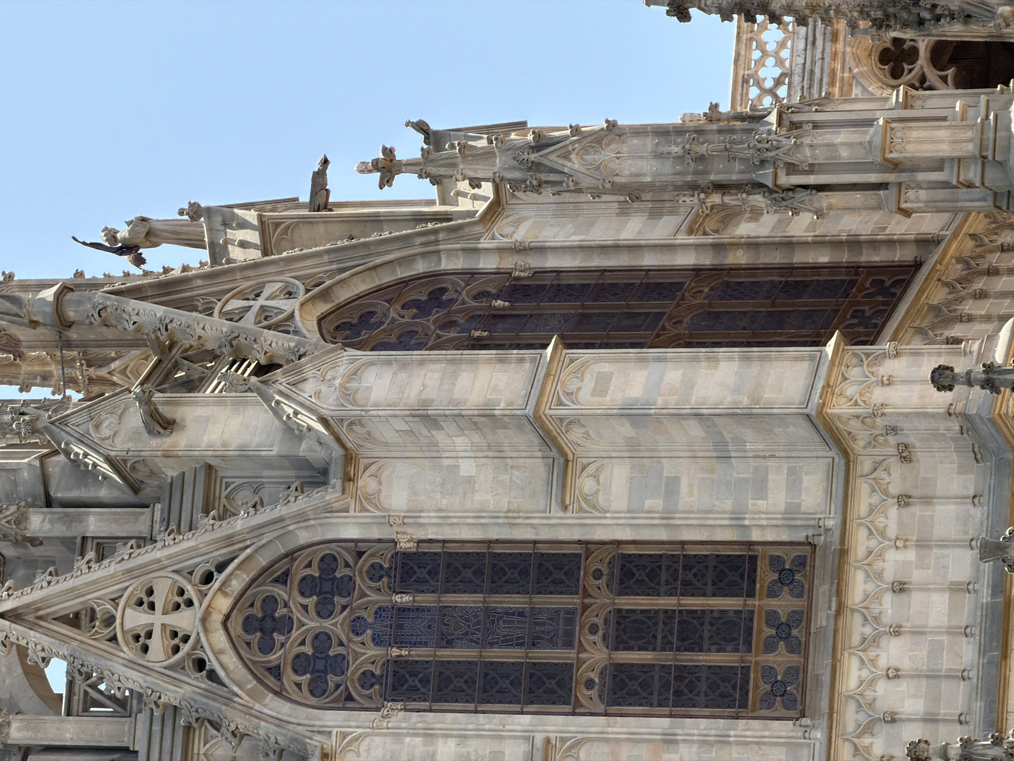 This is a medium-shot, eye-level, outdoor photograph of a section of a Gothic cathedral's exterior under a clear blue sky. The building is constructed of light-colored stone and features intricate architectural details, including ornate gargoyles, pointed arches, and stained-glass windows. The windows are large and rectangular, divided into many smaller panes by stone mullions. Some of the panes appear to have dark blue or black stained glass. The overall impression is one of grandeur and historical significance. There is no text visible in the image. The scene suggests it is daytime due to the bright sunlight casting shadows on the building's facade.