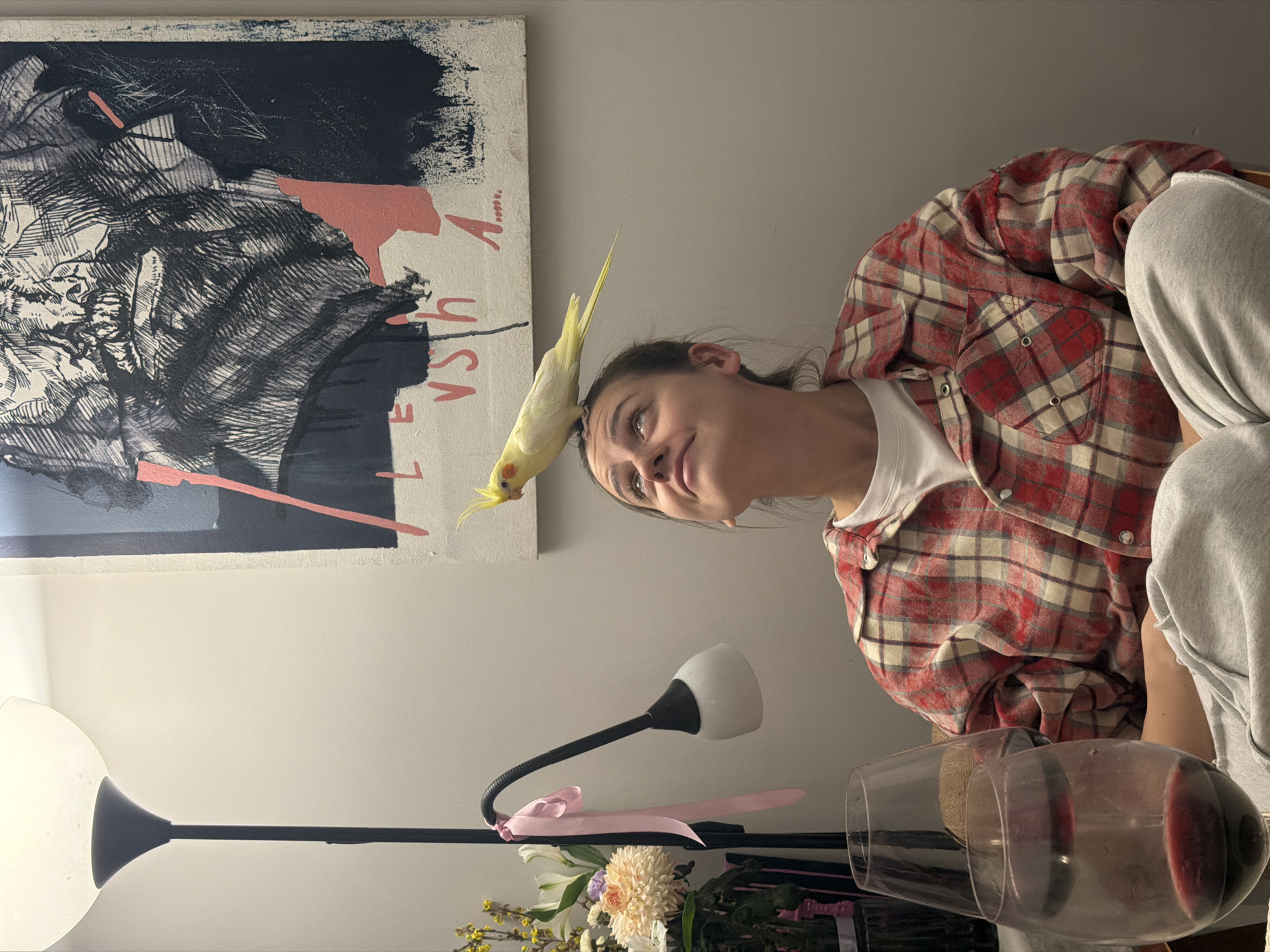 This indoor scene features a young woman interacting with a pet bird. The woman, shown from the chest up, has dark hair and a light complexion. She wears a red and white plaid flannel shirt over a white undershirt, with light grey sweatpants visible. Her expression is a soft smile or smirk as she looks upwards. A yellow cockatiel with an orange cheek patch is perched on her head, its crest and tail feathers pointing upwards.
The background wall is a light grey. A large abstract artwork hangs on the wall, featuring dark blue/black and white brushstrokes, intricate line work forming a figure, and splashes of light pink. Upside down, the text "LEVSYA" and a partial "A" are visible on the artwork.
To the left, a modern floor lamp with a glowing spherical white shade provides illumination. Below it, a black gooseneck desk lamp with a white cone shade is decorated with a pink ribbon. Fresh flowers, including white lilies, light orange chrysanthemums, and yellow sprigs, are arranged nearby. In the foreground, two clear glasses are visible, one containing a dark red liquid. The scene suggests a relaxed, domestic setting, likely under artificial lighting.