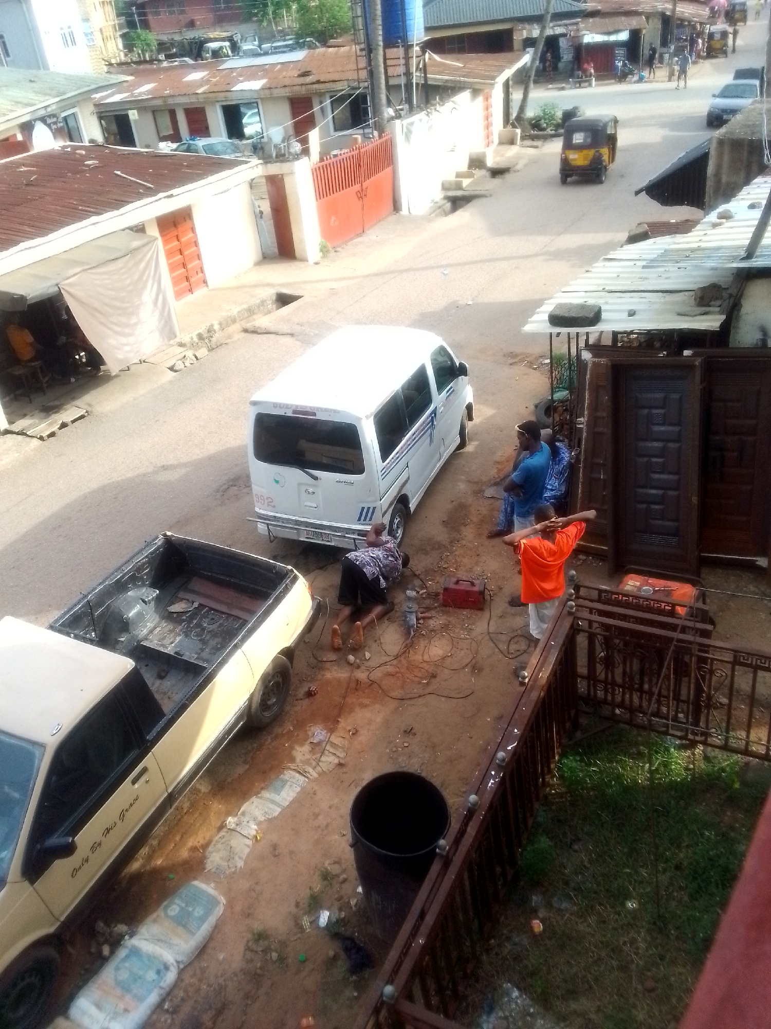The image shows a street scene in an urban area of Unknown, Unknown. A white van is parked on the side of the street near a yellow pickup truck with the words "Only By His Grace" on the side. A person is lying down next to the van, seemingly working on it, with tools nearby. Another person in an orange shirt is standing, looking towards the people standing near the doors. Three dark-colored wooden doors are propped up against a building. Two people stand nearby. A red welder is also visible. The street shows signs of foot traffic. Buildings with corrugated metal roofs and some with painted facades line the street. A yellow auto rickshaw is visible further down the road. A black bin sits near a fence. The setting appears to be a mix of residential and commercial, potentially a workshop or small business area.