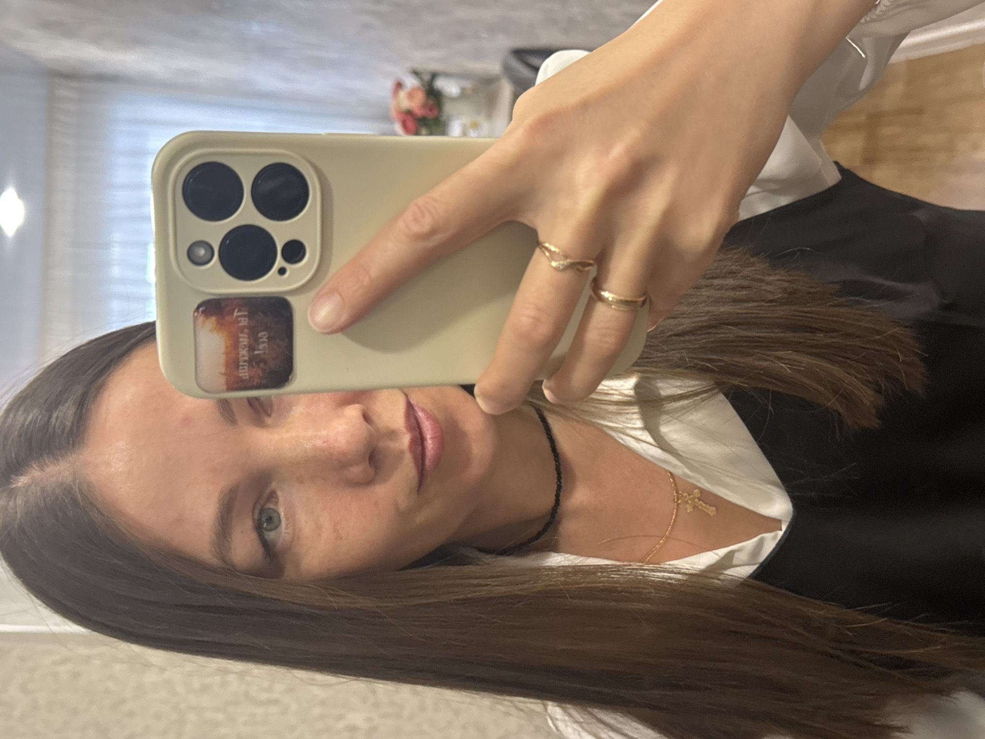 A selfie of a fair-skinned woman with brown hair and green eyes. She is wearing a white shirt and a black vest, with a gold cross necklace visible. She is holding a light-colored iPhone with a four-lens camera and a sticker on the back. The background is blurred, but curtains and a hint of flowers suggest an interior setting. The image was taken in Chaikovsky, Russia.