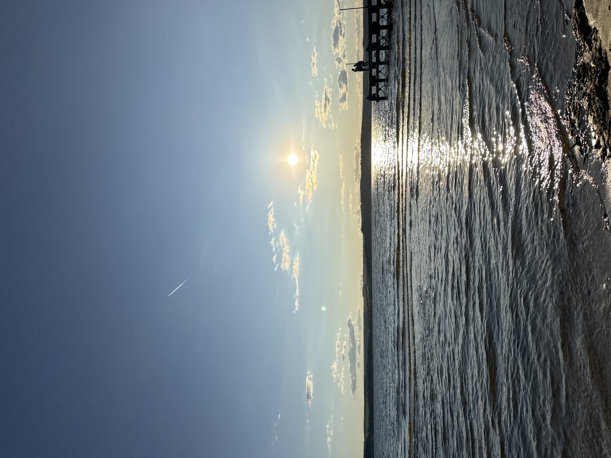 The image depicts a serene lakefront scene in Матюшино, Russia, during sunset. The sun, a bright orb, is low on the horizon, casting a warm golden glow that reflects shimmering on the rippling water. A thin contrail from an airplane stretches across the vast blue sky, adding a touch of human presence to the natural landscape. On the right side of the frame, a wooden pier extends into the lake, with two silhouetted figures standing at its end, appearing to be in conversation or admiring the view. The shoreline in the foreground is composed of sand and some scattered debris. The background features a distant, tree-lined horizon under a sky dotted with a few fluffy white clouds. The overall atmosphere is tranquil and picturesque.